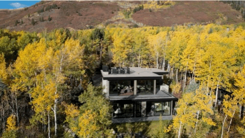 Modern house surrounded by yellow fall trees in a forested area with hills in the background.