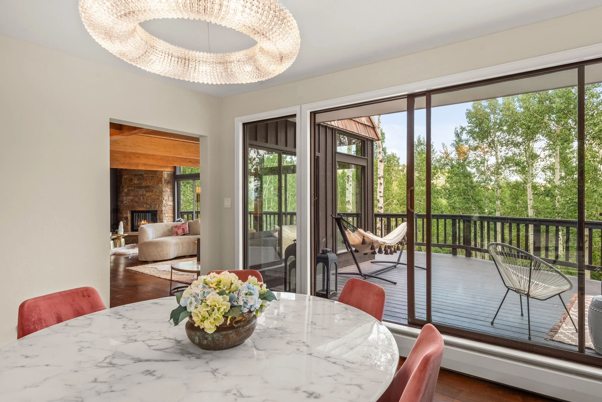 Indoor dining area with a white marble table and pink chairs, and an outdoor balcony with a hammock and seating, overlooking trees.