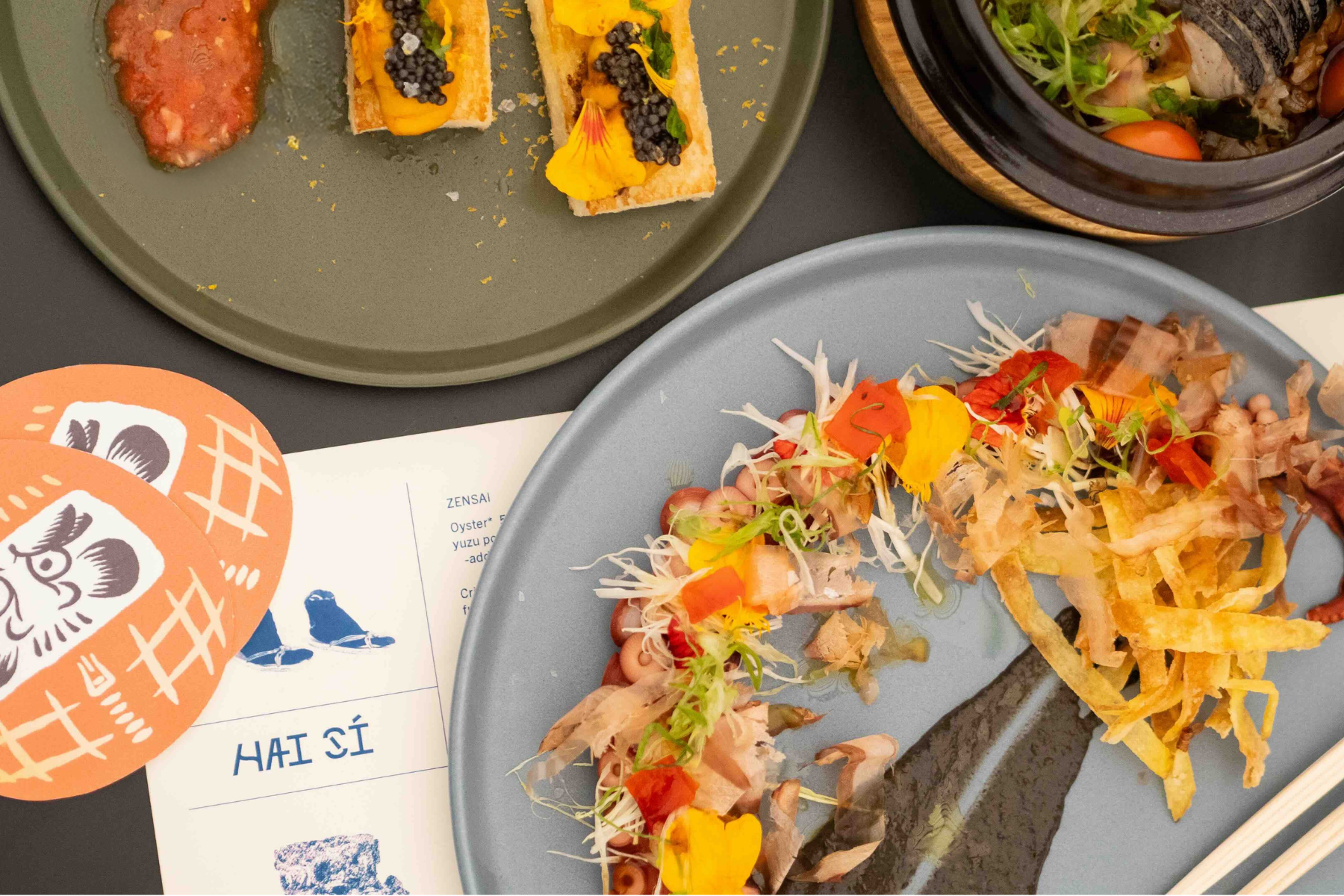 Assorted Asian dishes on plates, including sushi rolls with flowers and garnishes, and a bowl of ramen with vegetables, all presented on a dark table.