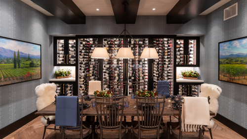 A dining room with a wooden table set for six, surrounded by chairs with various upholstery, some with sheepskin throws. Above the table hangs a chandelier with three shades. Behind the table, a wine storage wall holds numerous bottles of wine, flanked on either side by small shelves with potted plants. The room features two landscape paintings on textured gray wallpaper and a hardwood floor.
