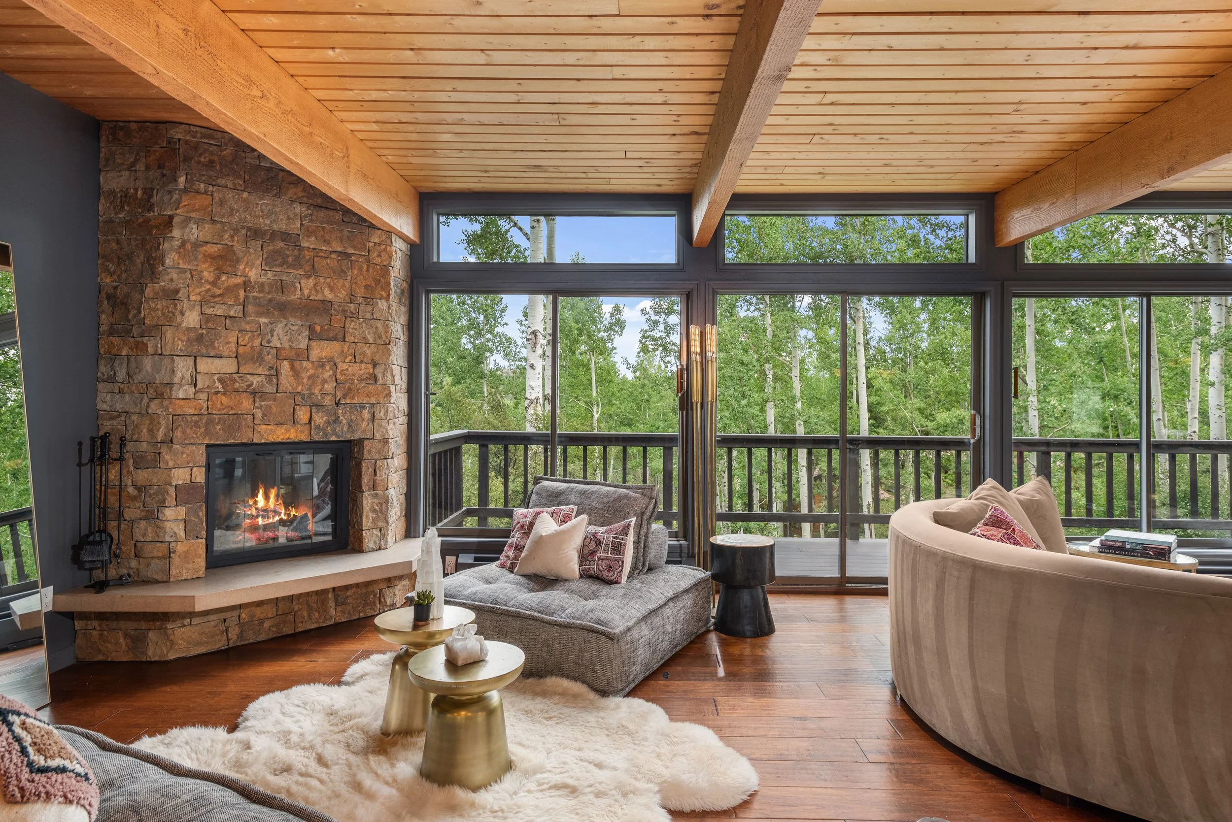 Cozy living room with a stone fireplace, large windows showing a wooded view, and comfortable furniture including a gray lounge chair, a curved beige sofa with pillows, and gold-colored side tables on a white shag rug.