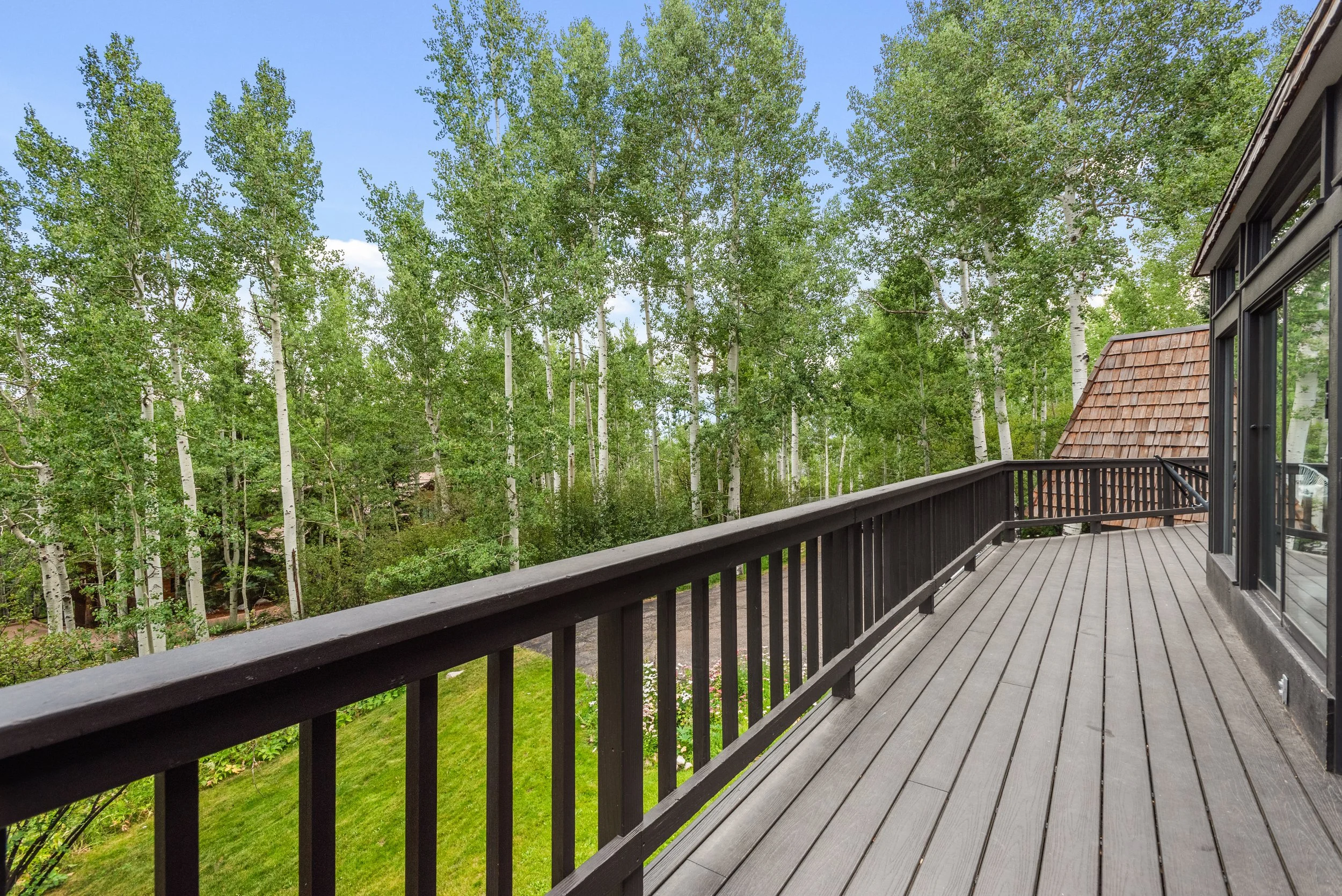 A spacious outdoor wooden deck with black railing overlooking a green backyard with tall trees and a partly cloudy sky.