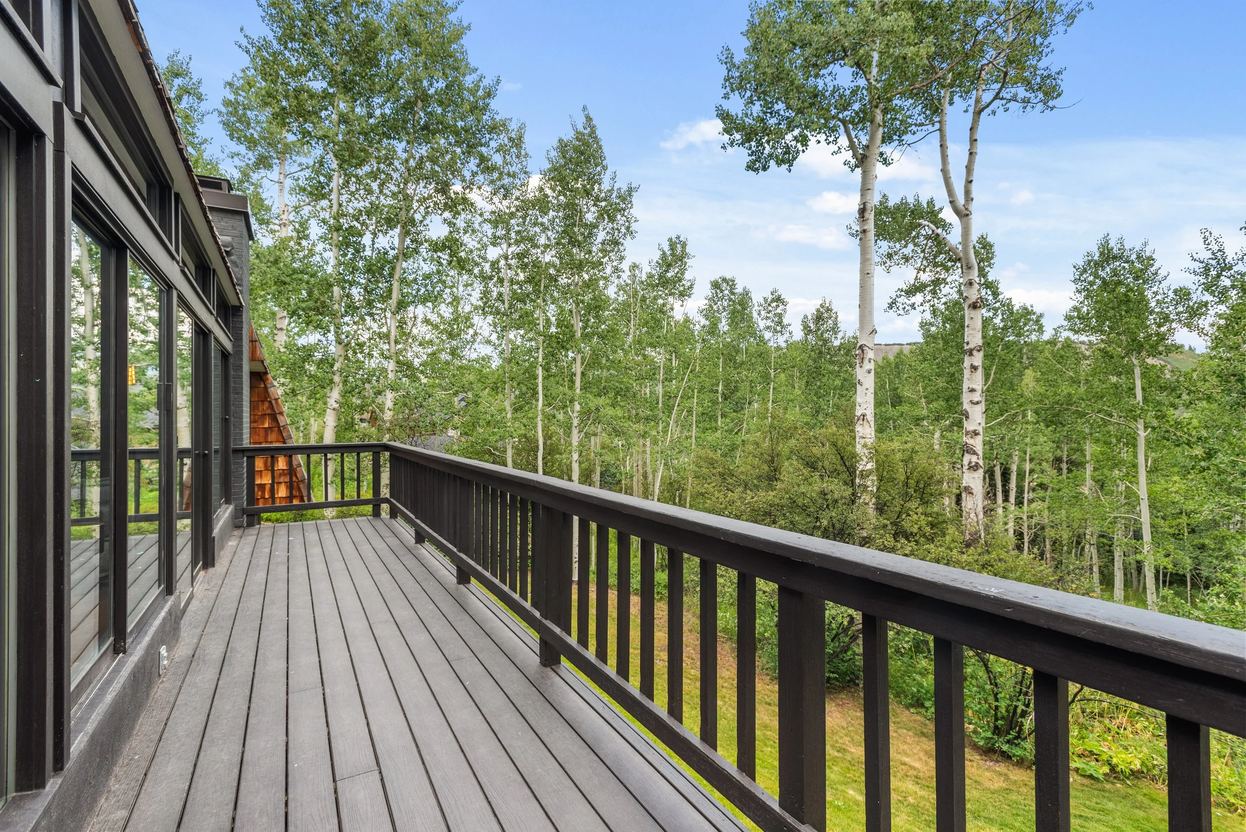 Empty balcony with wooden floor and black railing overlooking green trees and a partly cloudy sky.