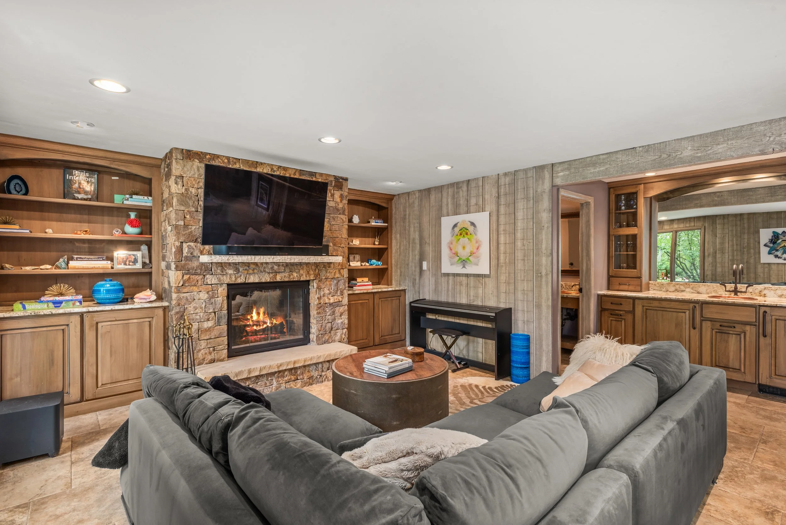 Living room with a gray sectional sofa, a round coffee table, a fireplace, a mounted television, built-in wooden shelves, and a view into a kitchen.