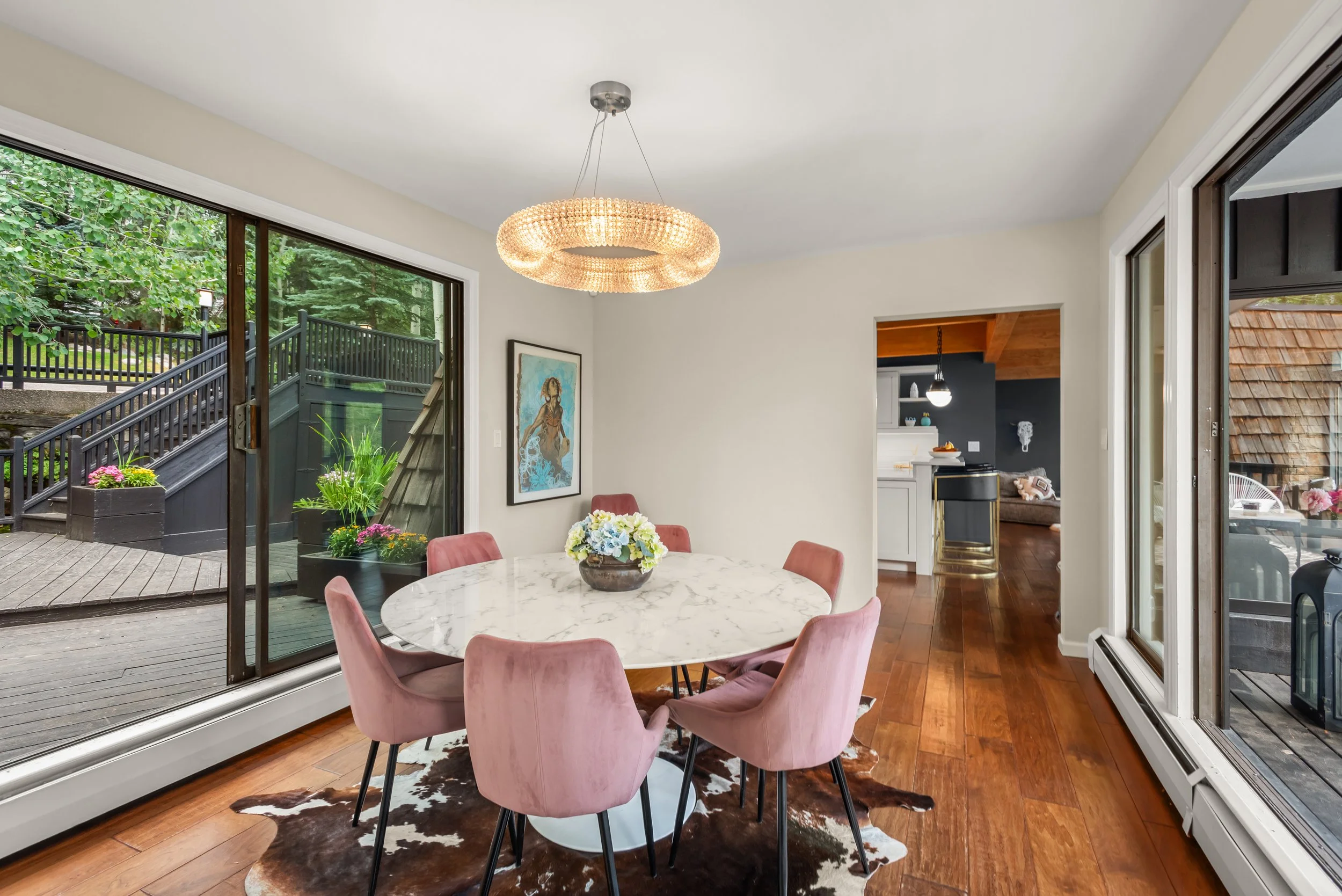 Dining room with a round marble table surrounded by six pink velvet chairs, a cowhide rug underneath, a floral centerpiece, large sliding glass doors leading to an outdoor deck, a chandelier overhead, and an open doorway to the kitchen in the backgro