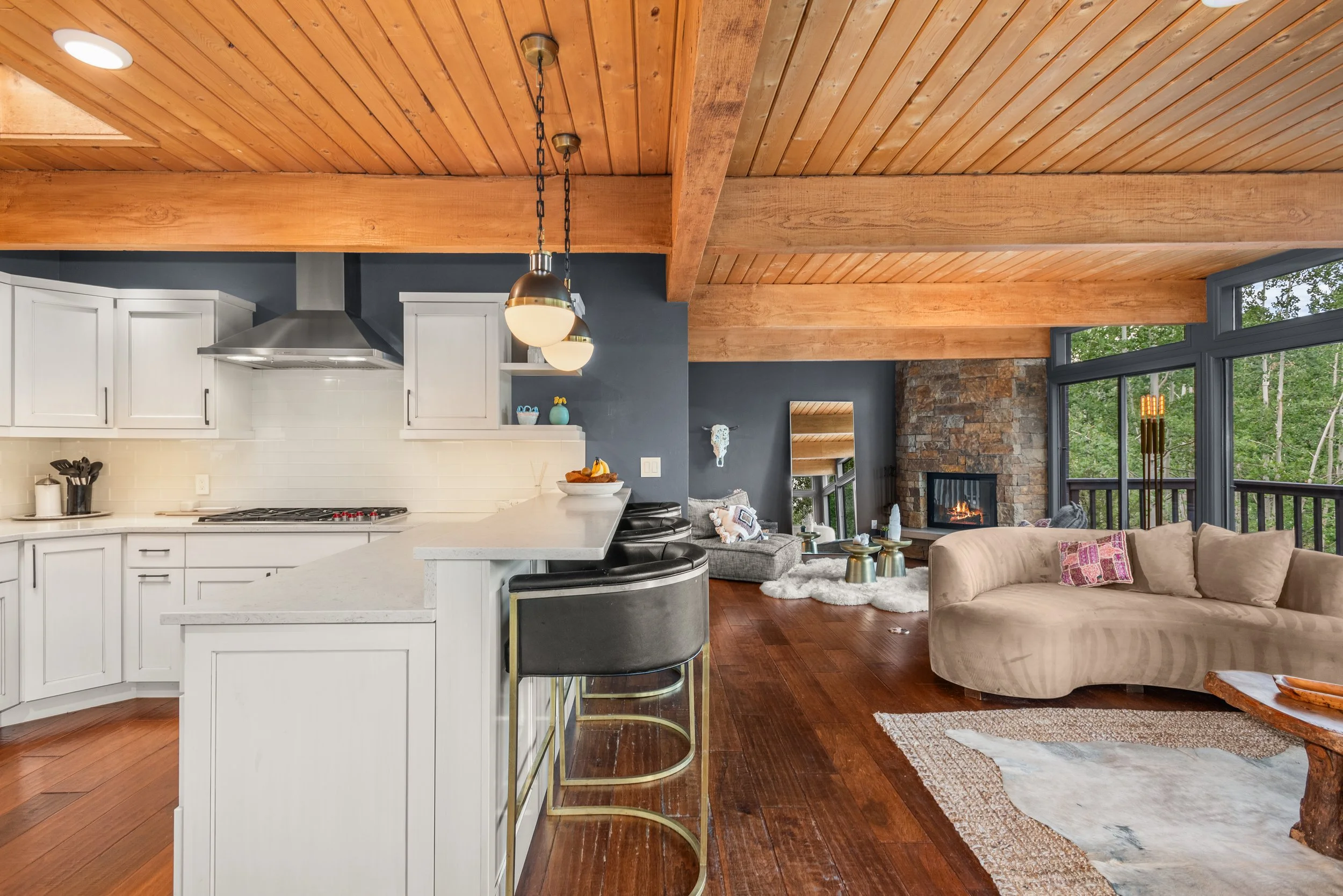 Open-concept living room and kitchen with wooden ceiling beams, white cabinetry, gray accent wall, stone fireplace, large windows overlooking a wooded area, beige sofa, and modern decor.