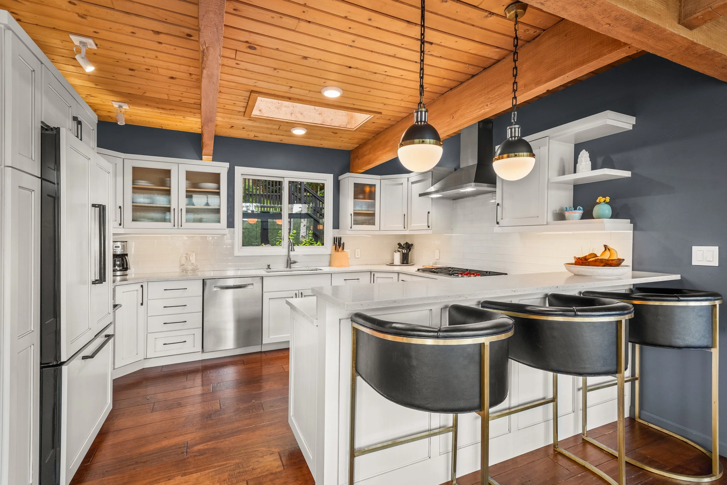 Modern kitchen with white cabinets, a white countertop, stainless steel appliances, pendant lights, and a wooden ceiling with exposed beams.