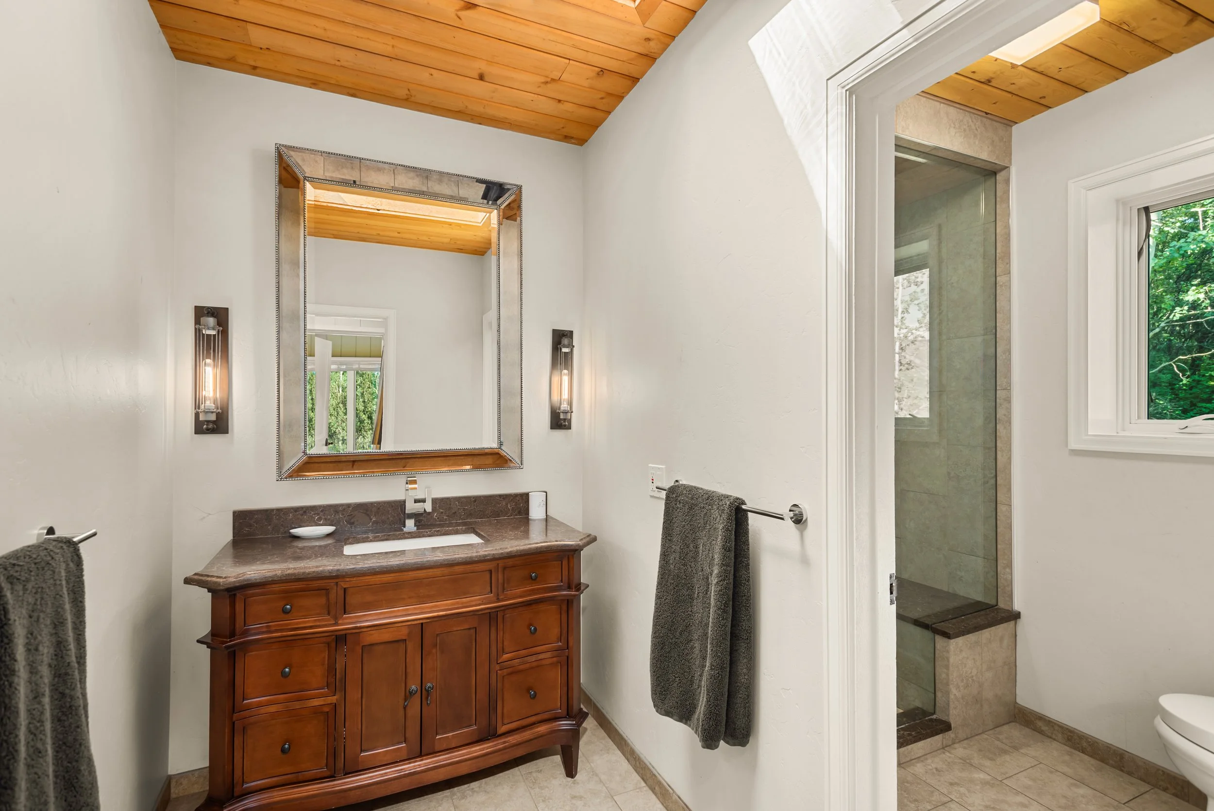 Bathroom vanity with wooden cabinet, large mirror, and two wall-mounted lights. To the right, a walk-in shower with tiled walls and bench, next to a window showing green trees outside.