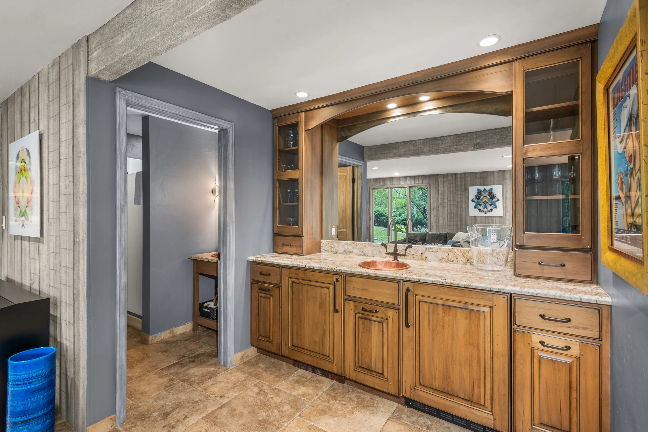 Kitchen area with wooden cabinets, granite countertops, a small copper sink, and a pass-through window to the living room with large windows and greenery outside.