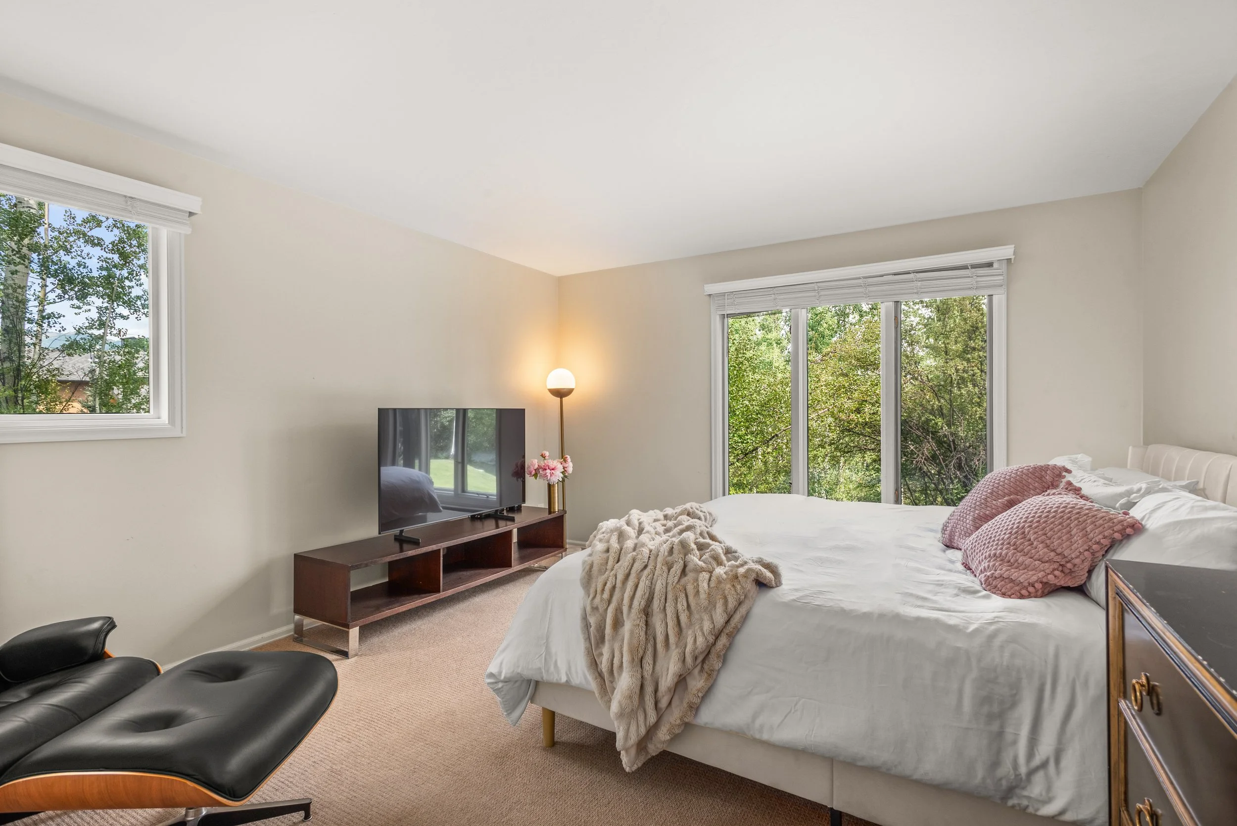 Bright bedroom with large window and sliding glass door, white bed with pink pillows, beige blanket, flat-screen TV on wooden stand, black lounge chair, and a standing lamp with a floral decoration.
