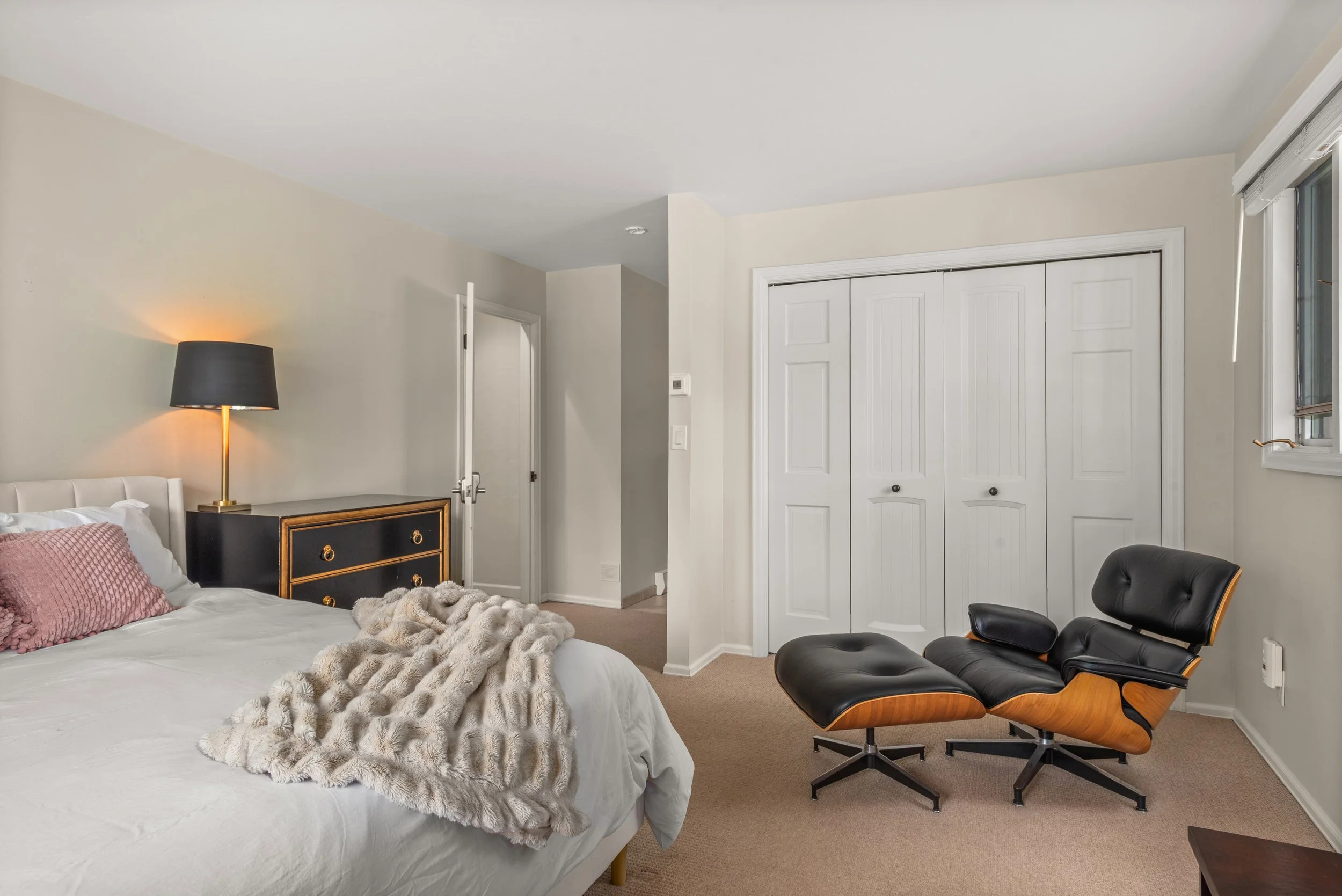 A bedroom with a bed, black dresser with gold accents, bedside lamp, black and wood lounge chair, and closed closet doors.