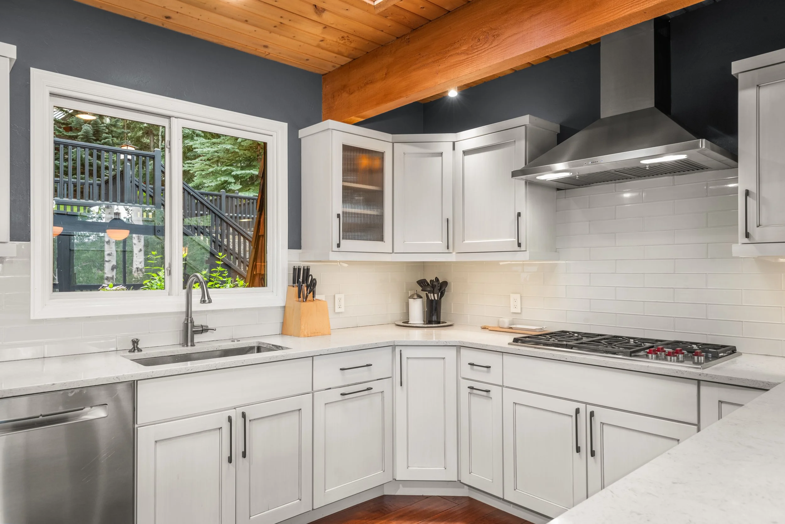 Modern kitchen with white cabinets, stainless steel dishwasher, gas stove, stainless steel range hood, and white subway tile backsplash, overlooking a backyard with trees and a black staircase.