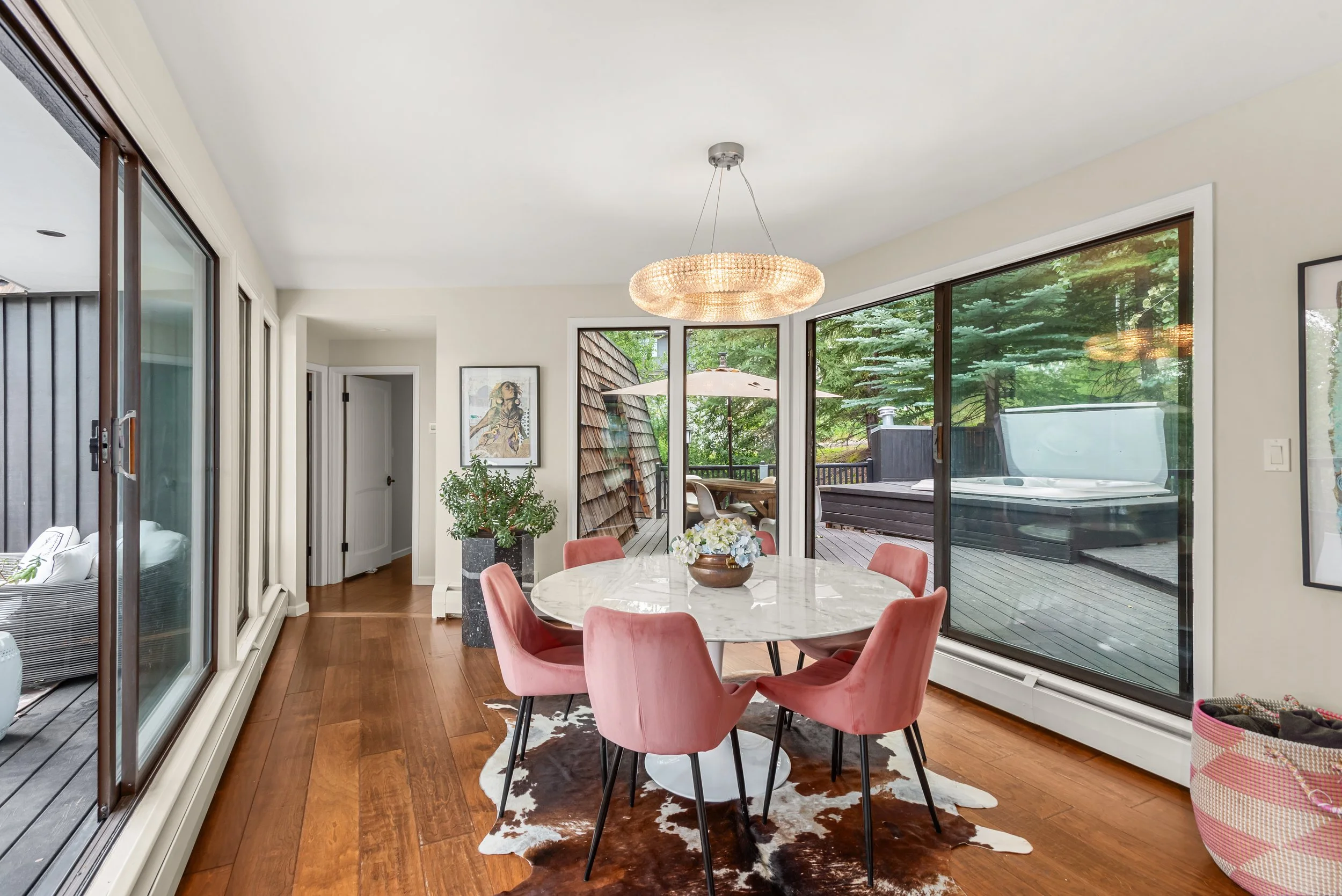 Dining room with pink chairs around a marble table, sliding glass doors to a deck, and large windows showing greenery outside.