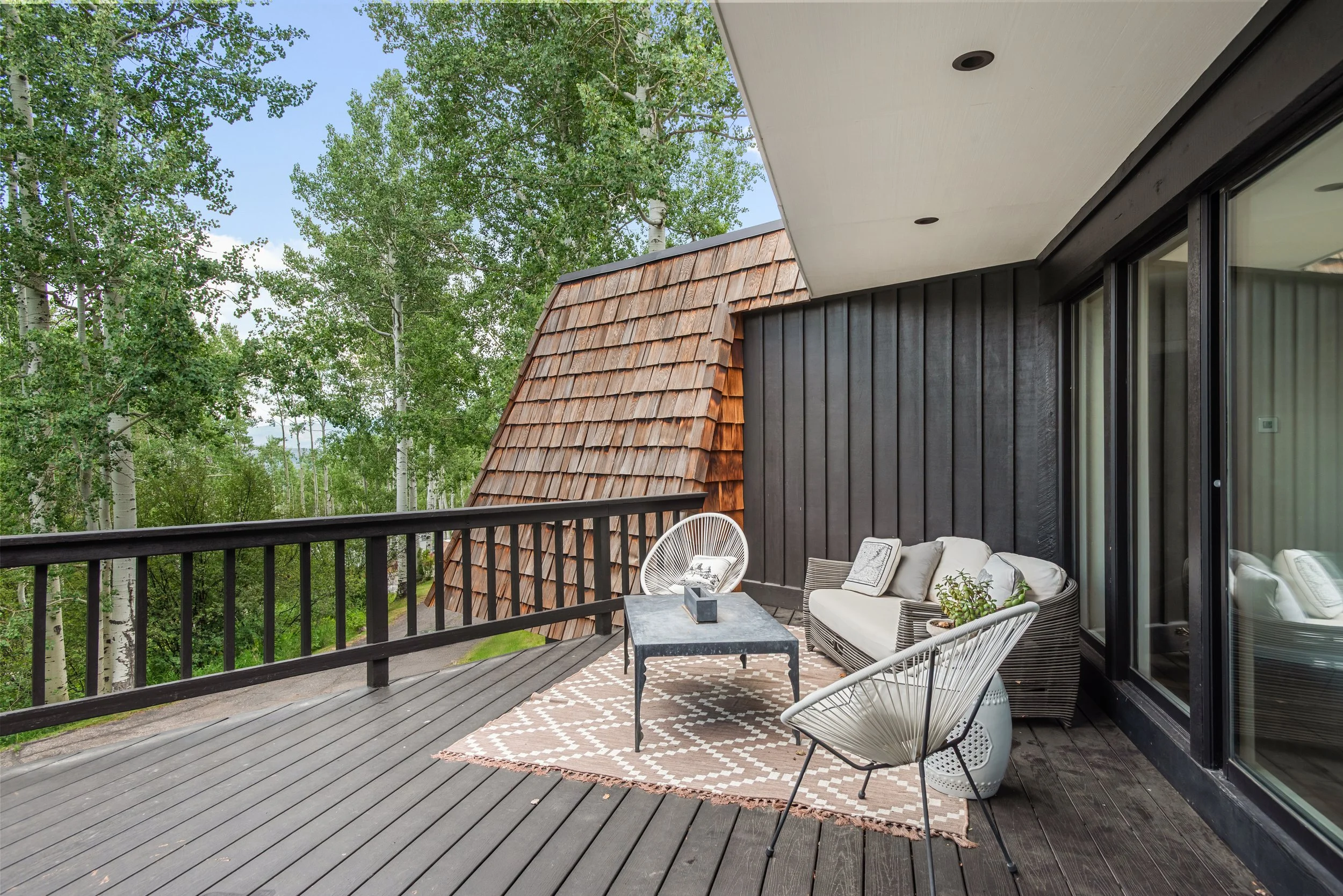 Outdoor balcony with black wooden railing, beige seating, black coffee table, and patterned outdoor rug, surrounded by green trees.