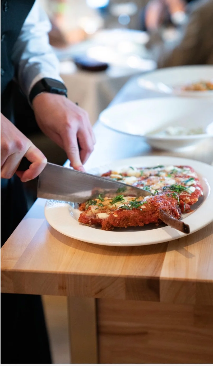 Person slicing a cooked meat dish, likely a chop, on a white plate at a dining table.