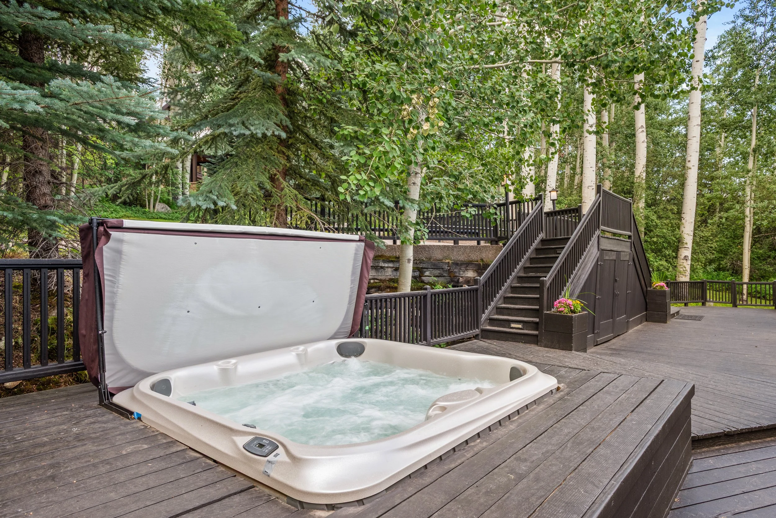 A hot tub on a wooden deck in a forested area with trees and a black staircase.