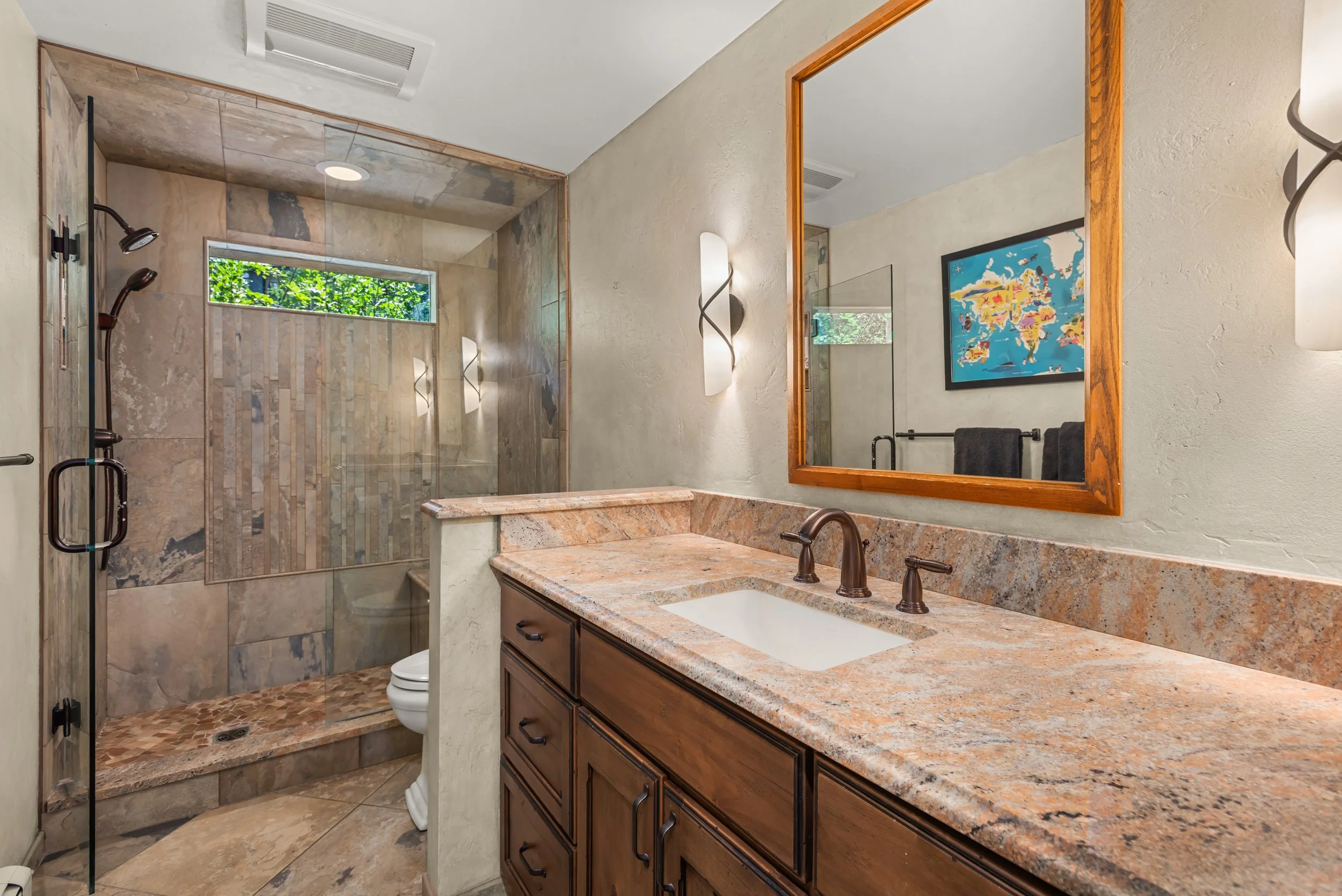 Bathroom with a granite countertop sink, wooden cabinet, framed mirror, and a walk-in shower with a small window and wall-mounted sconces.