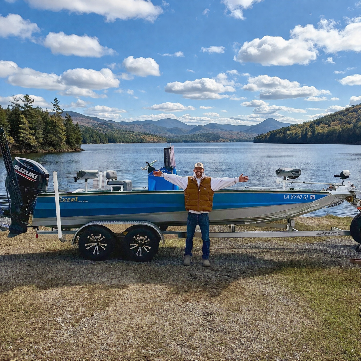 A small Tracker boat on a trailer parked near a body of water with hills and trees in the background and a partly cloudy sky.