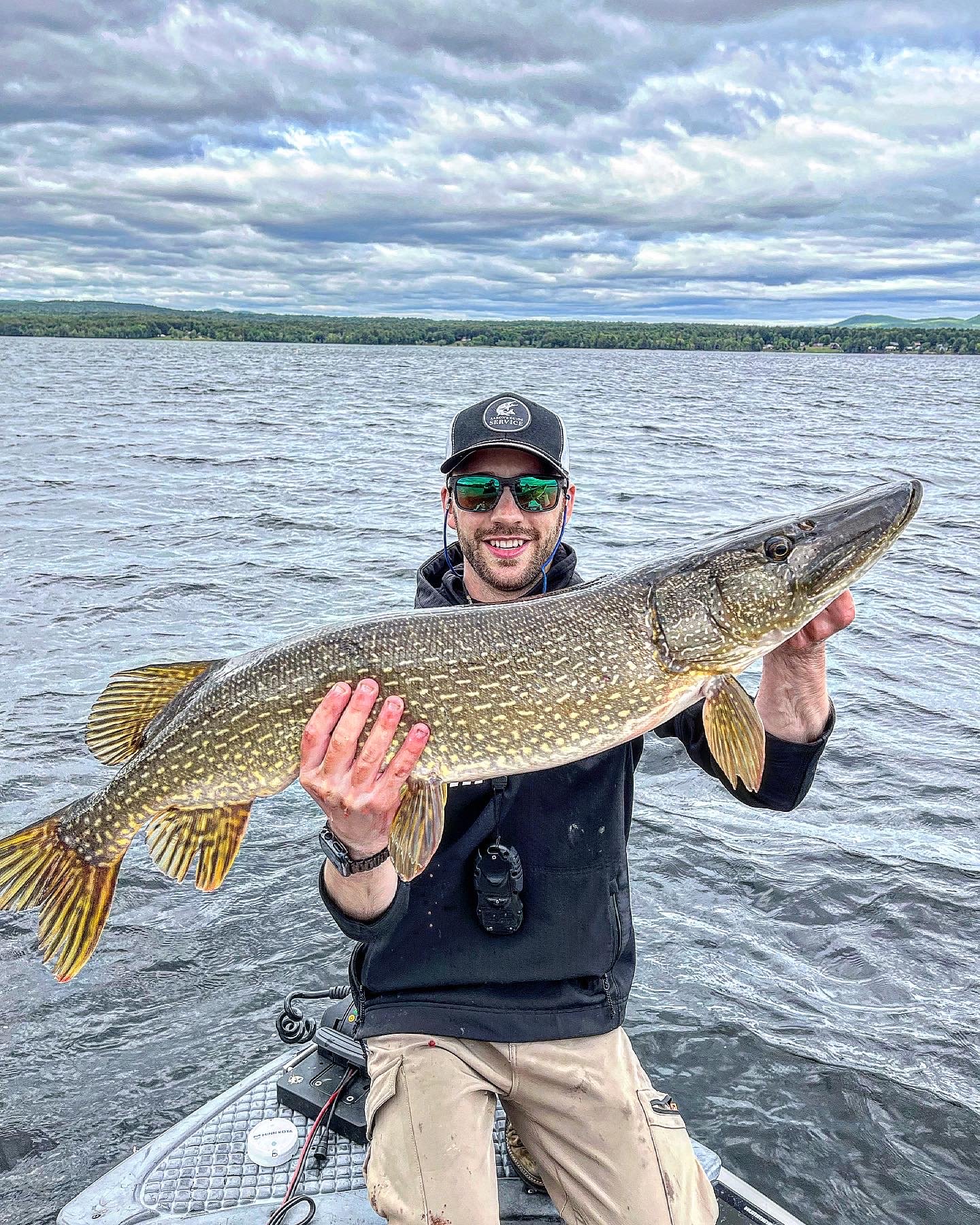 A man wearing sunglasses and a cap is smiling and holding a large fish, likely a pike, on a boat in a lake under a cloudy sky.
