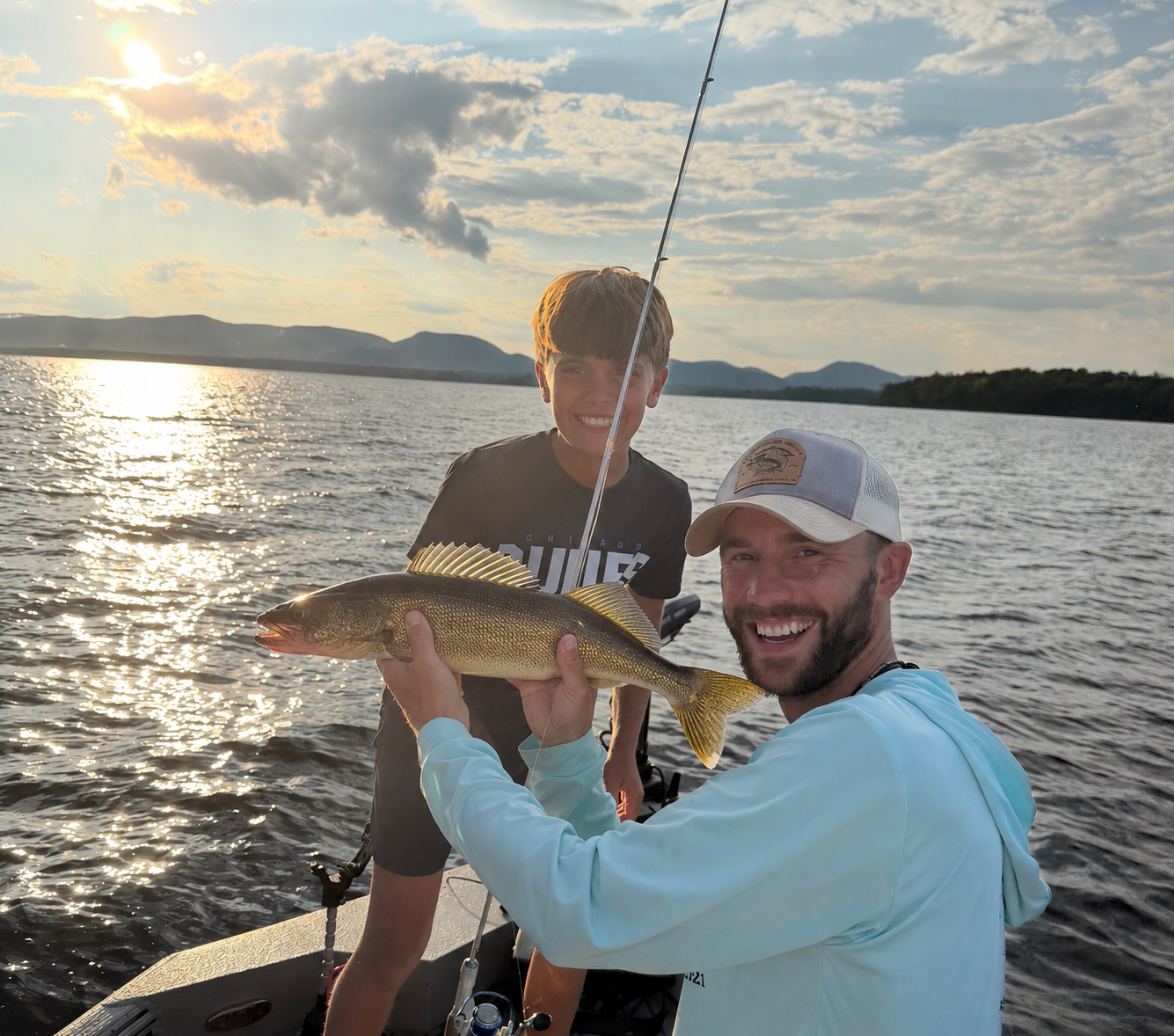 Two young men fishing from a boat on a lake under a partly cloudy sky, with trees and hills in the distance. One man is pointing toward the water while the other holds a fishing rod.