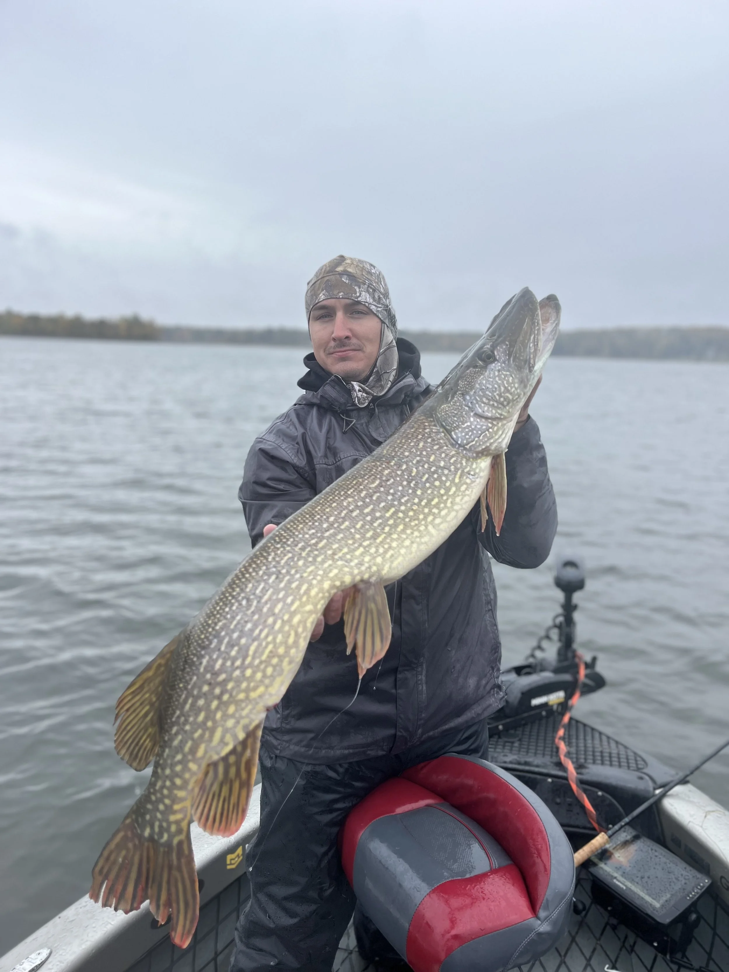 Man in rain gear on a boat holding a large fish, likely a northern pike, in a lake with cloudy sky.