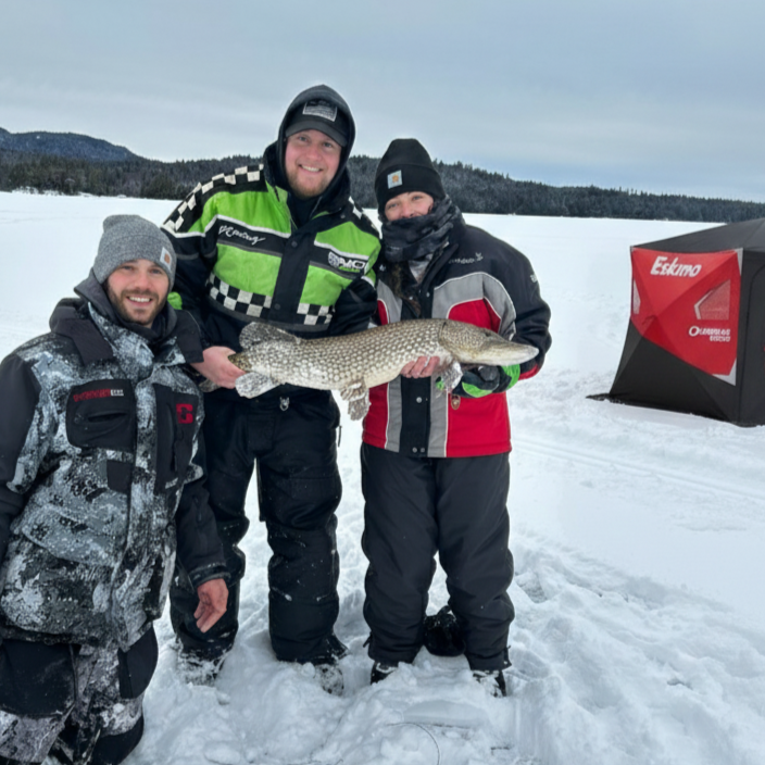 Man standing on snow-covered ice holding a large fish outdoors with trees in the background.