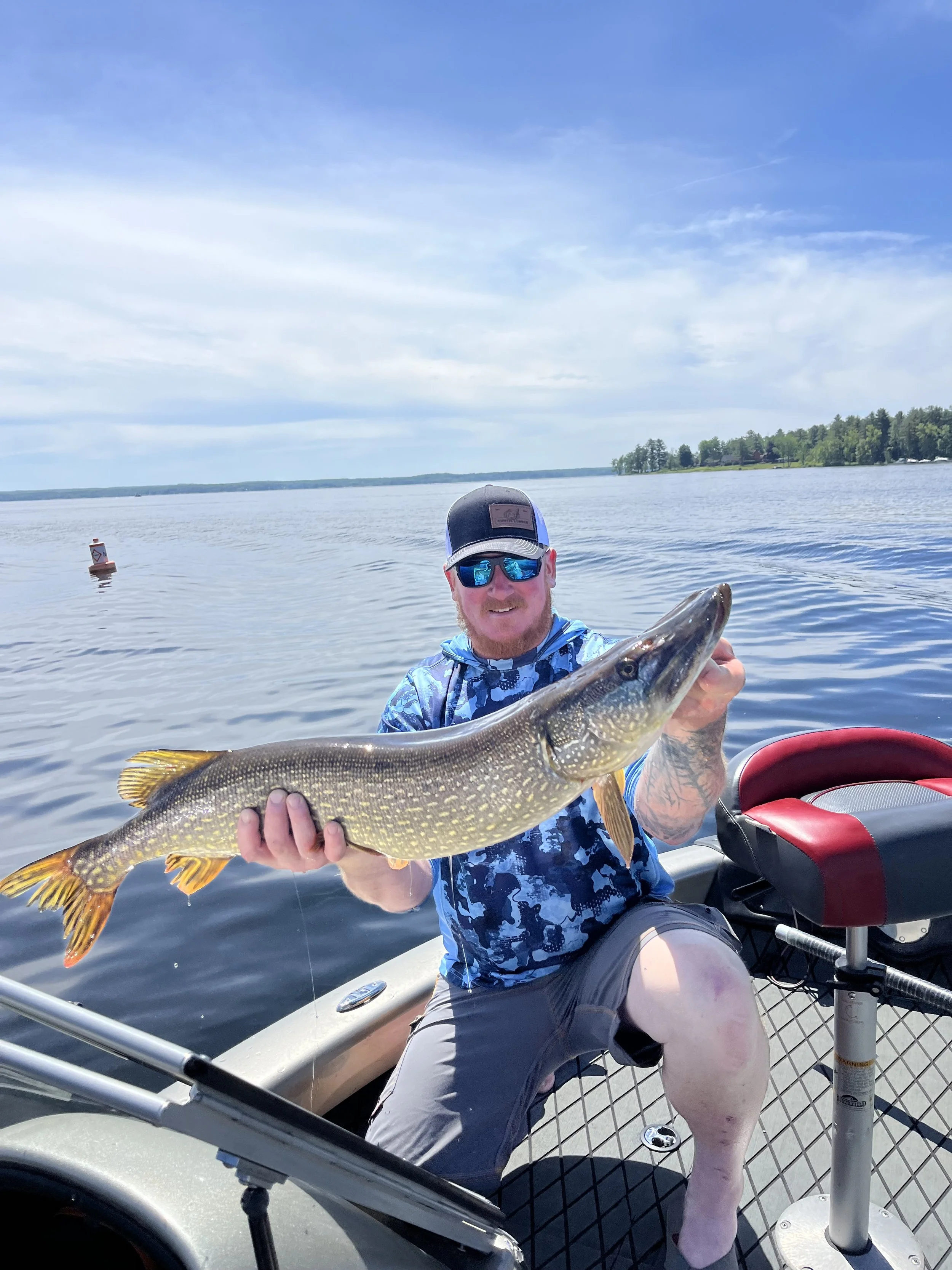 Man on a boat holding a large fish, wearing a cap, sunglasses, and a blue camouflage shirt, on a lake with trees in the background.