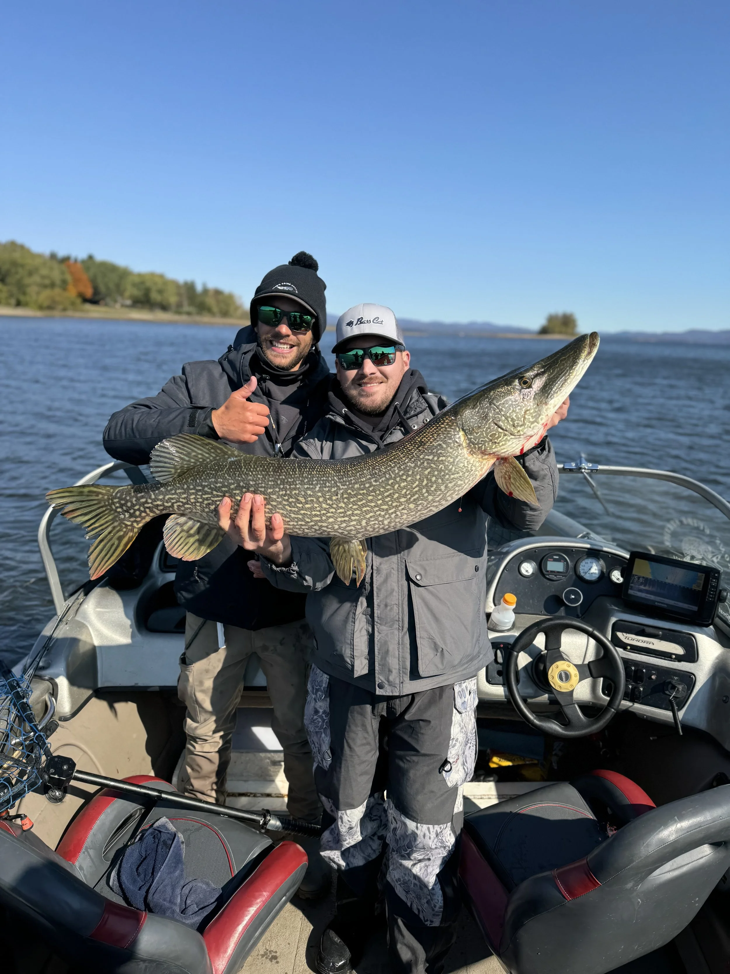 Two men on a boat holding a large fish, possibly a pike, with a blue sky and water in the background.