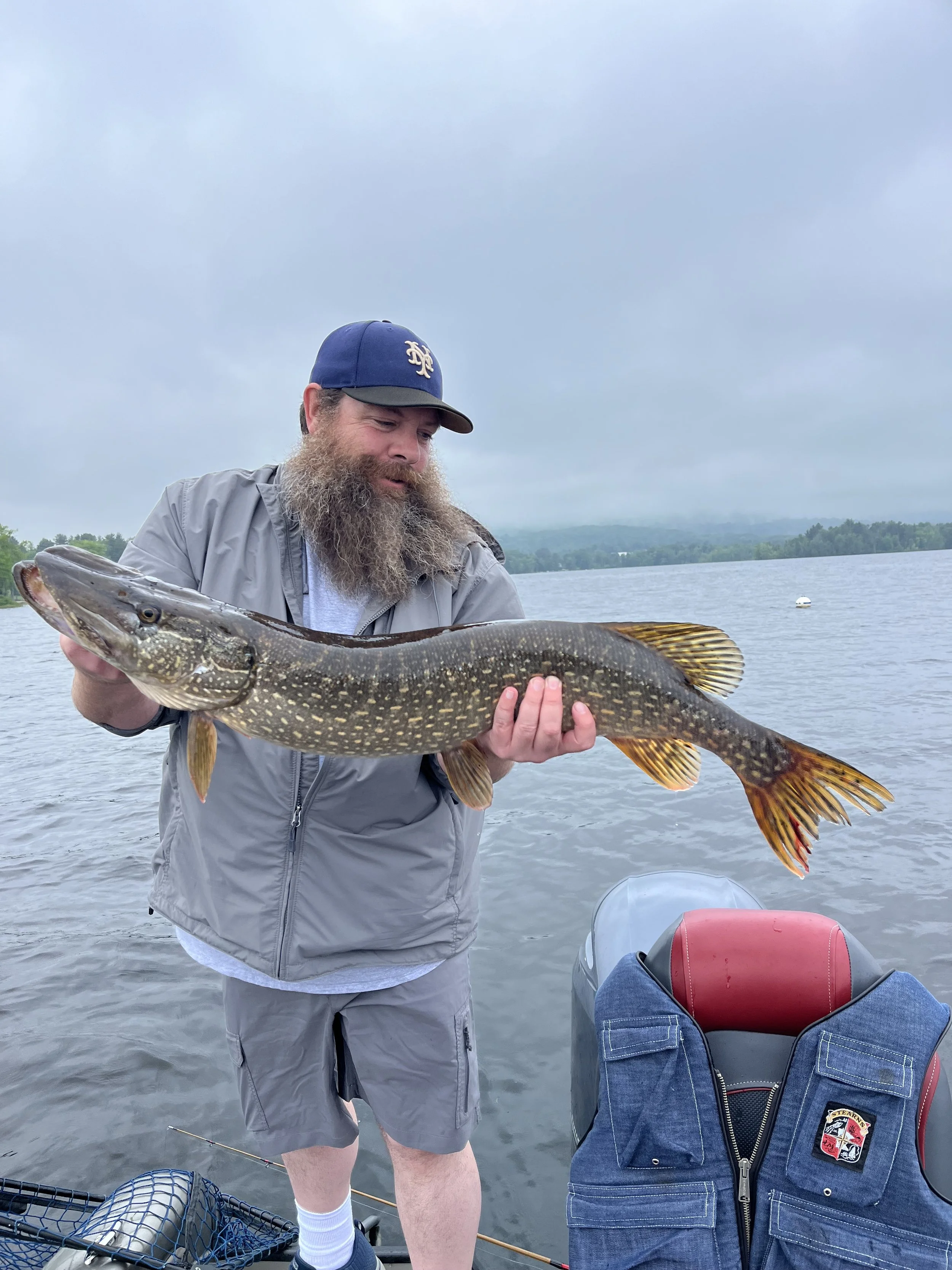 Man with a long beard, wearing a blue cap and gray jacket, holding a large fish with both hands on a boat, with a lake and cloudy sky in the background.