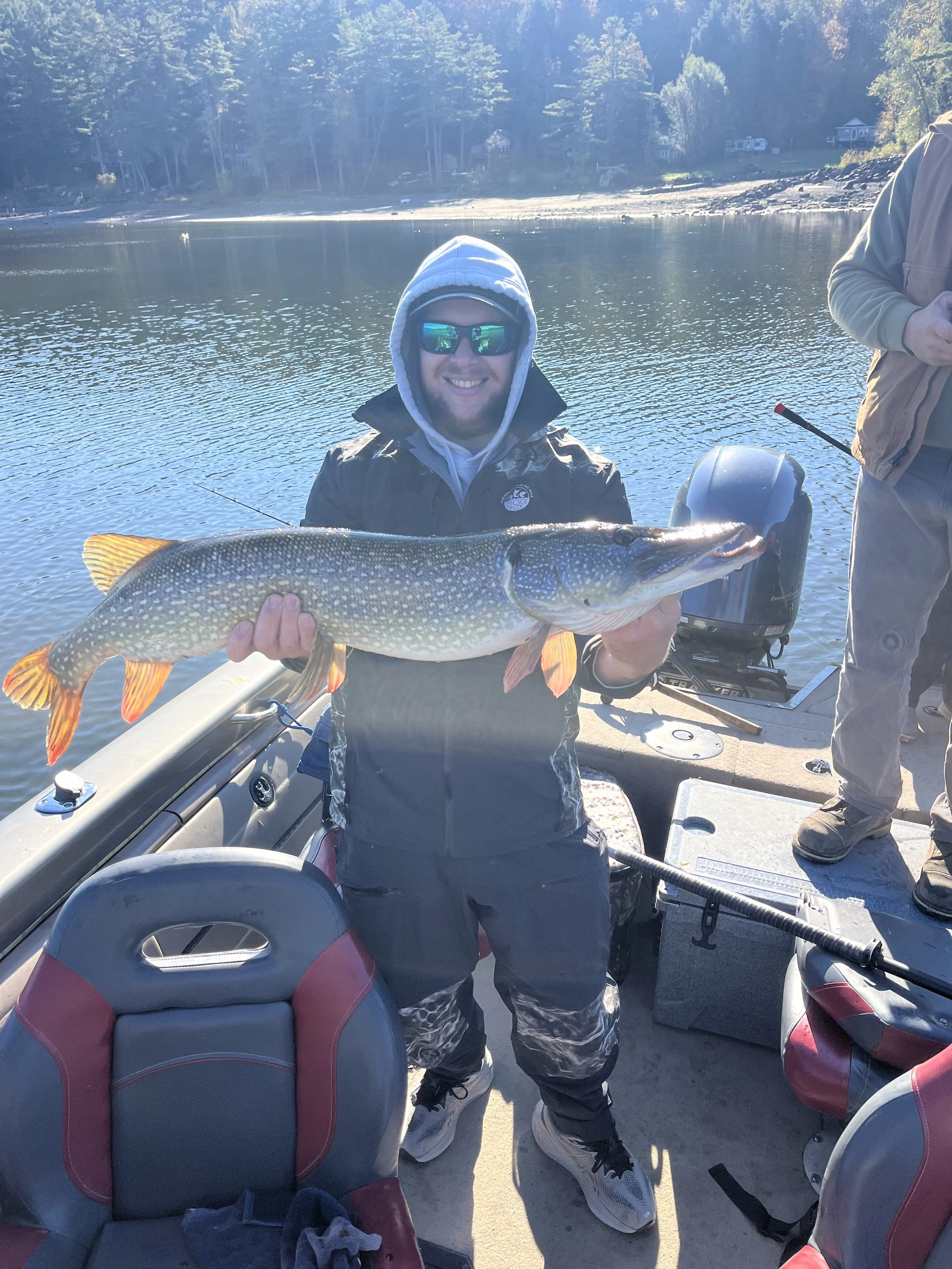 Man in sunglasses and a hoodie holding a large fish on a boat by a lake with forested shoreline in the background.