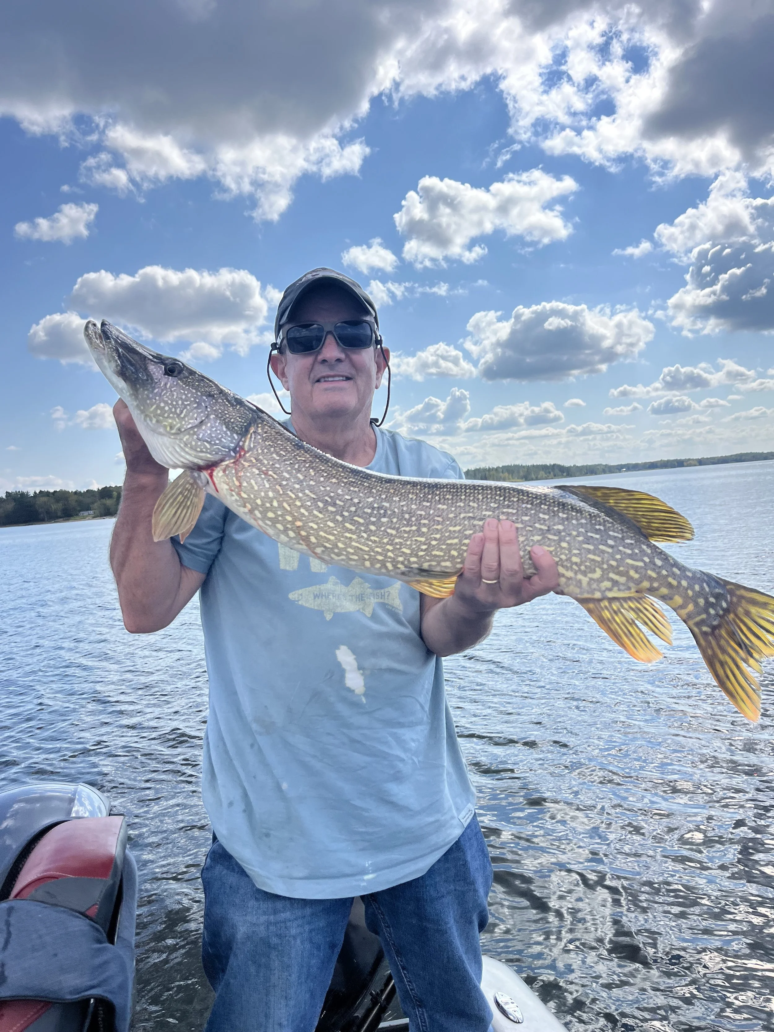 A man wearing sunglasses, a cap, and a gray T-shirt holding a large fish over water on a boat with a partly cloudy sky in the background.