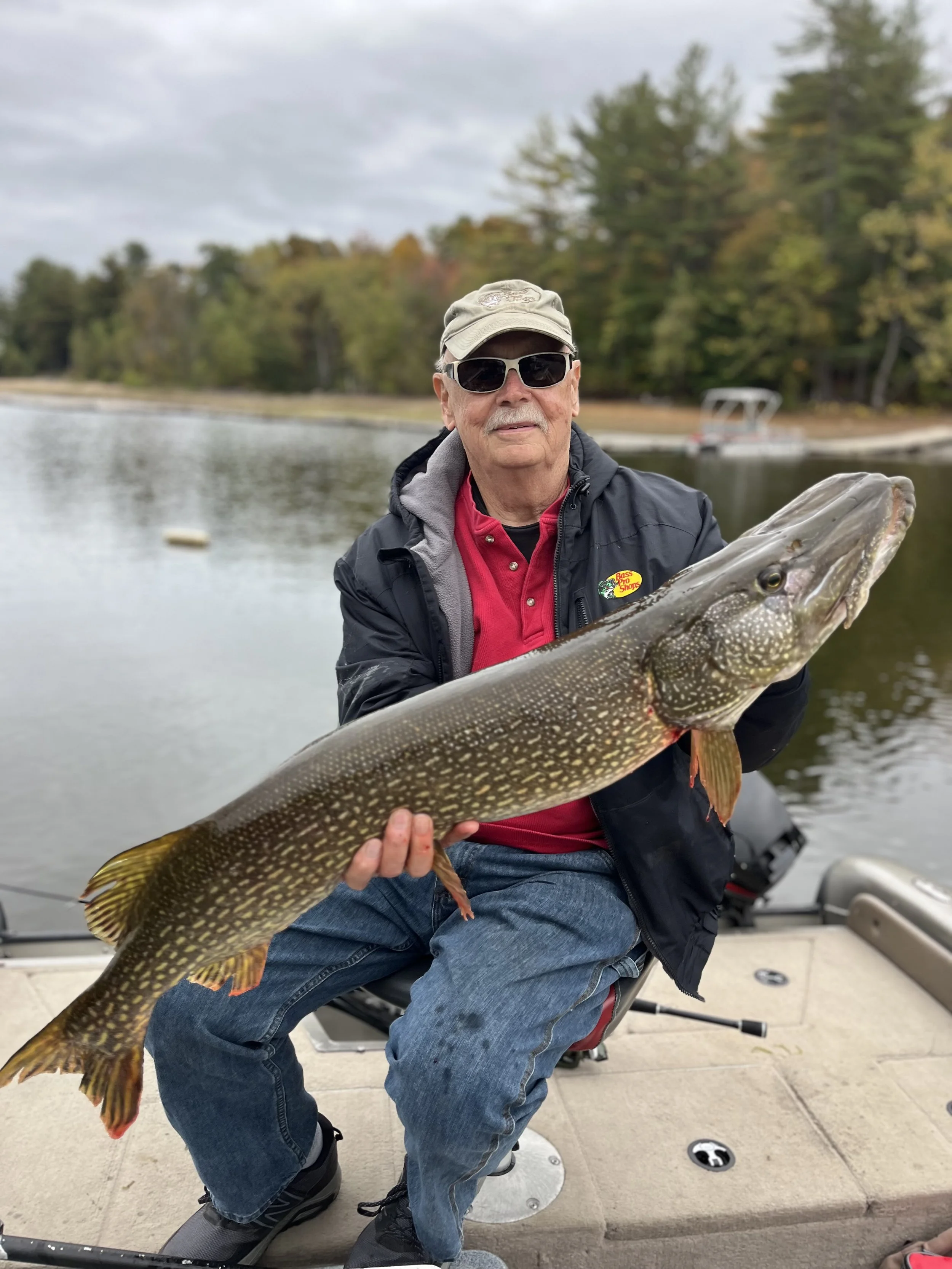 An older man wearing sunglasses, a hat, and a jacket is sitting on a boat, holding a large fish he caught, with water and trees in the background.
