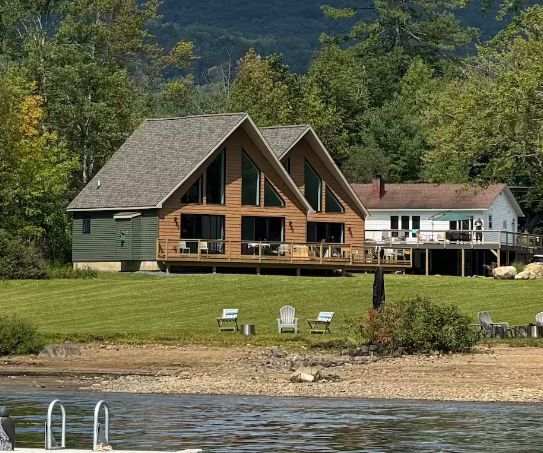 Living room with large triangular windows overlooking a grassy yard and lake, with a wooden deck, a turquoise armchair, a small side table, and two paddles hanging on the wall.