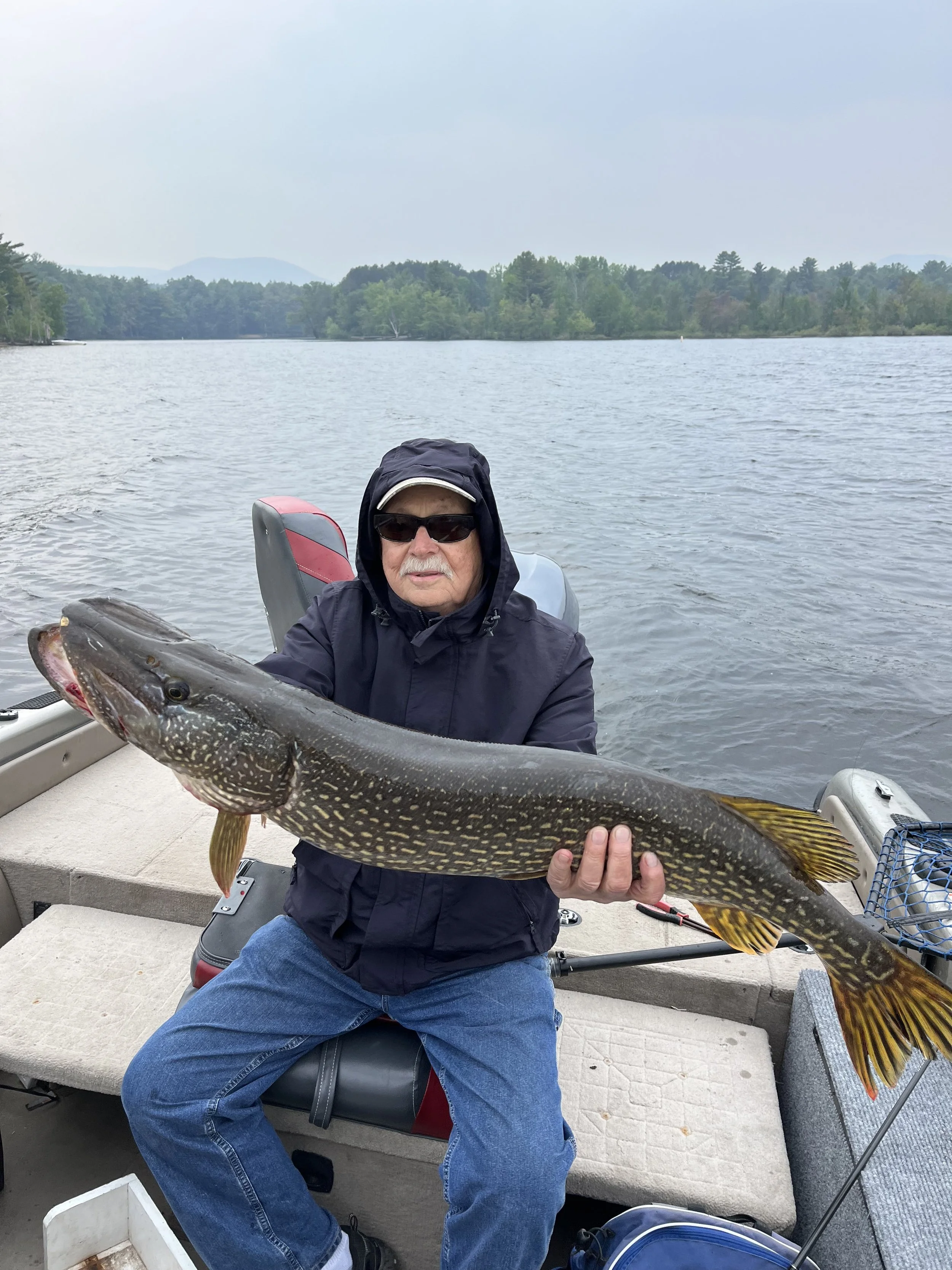 An elderly man wearing sunglasses, a black jacket with the hood up, and blue jeans, is sitting on a boat holding a large fish, likely a pike, across his lap. The boat is on a lake with trees and hills in the background.