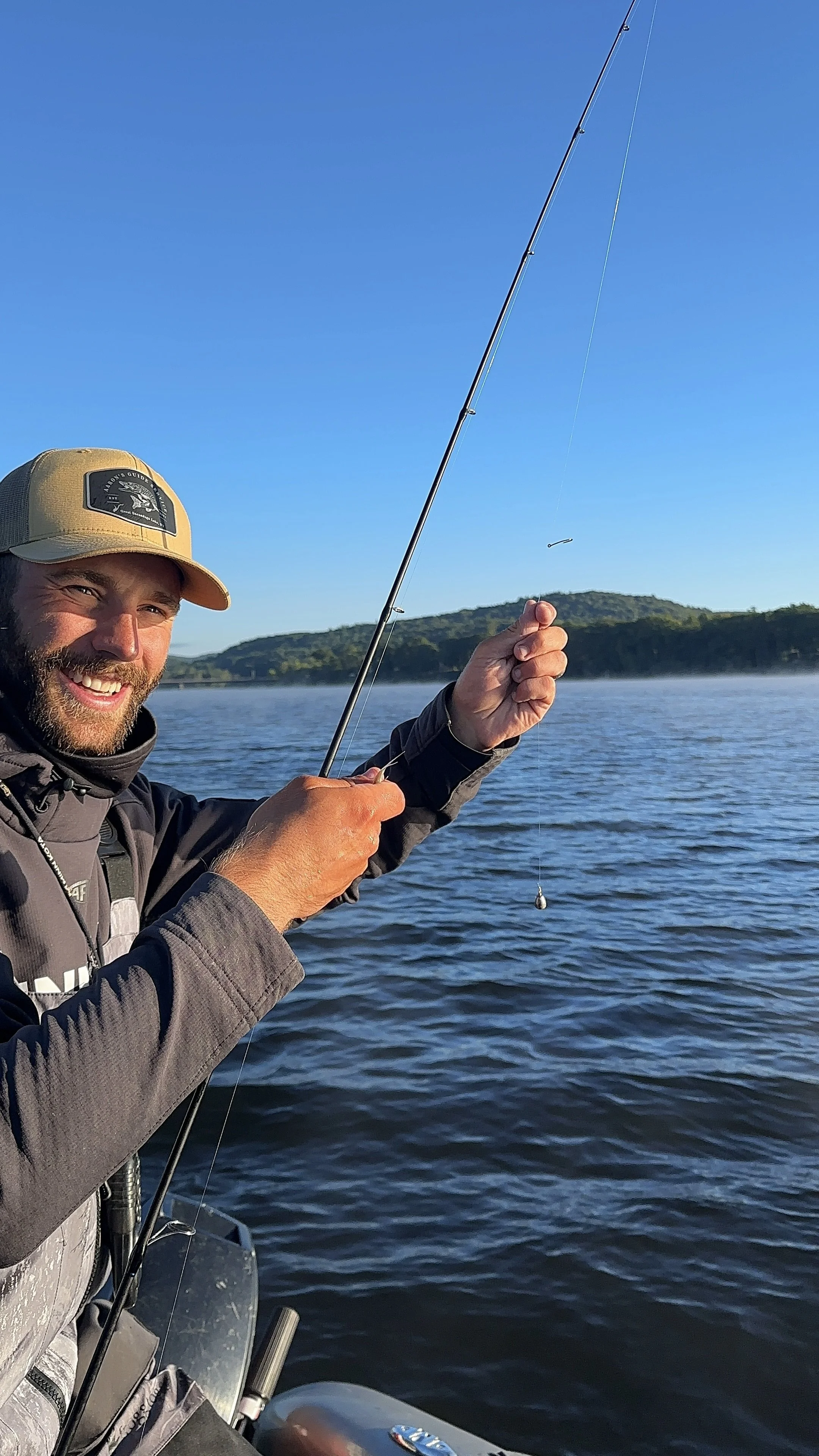 A man wearing sunglasses and a cap is smiling and holding a large fish, likely a pike, on a boat in a lake under a cloudy sky.