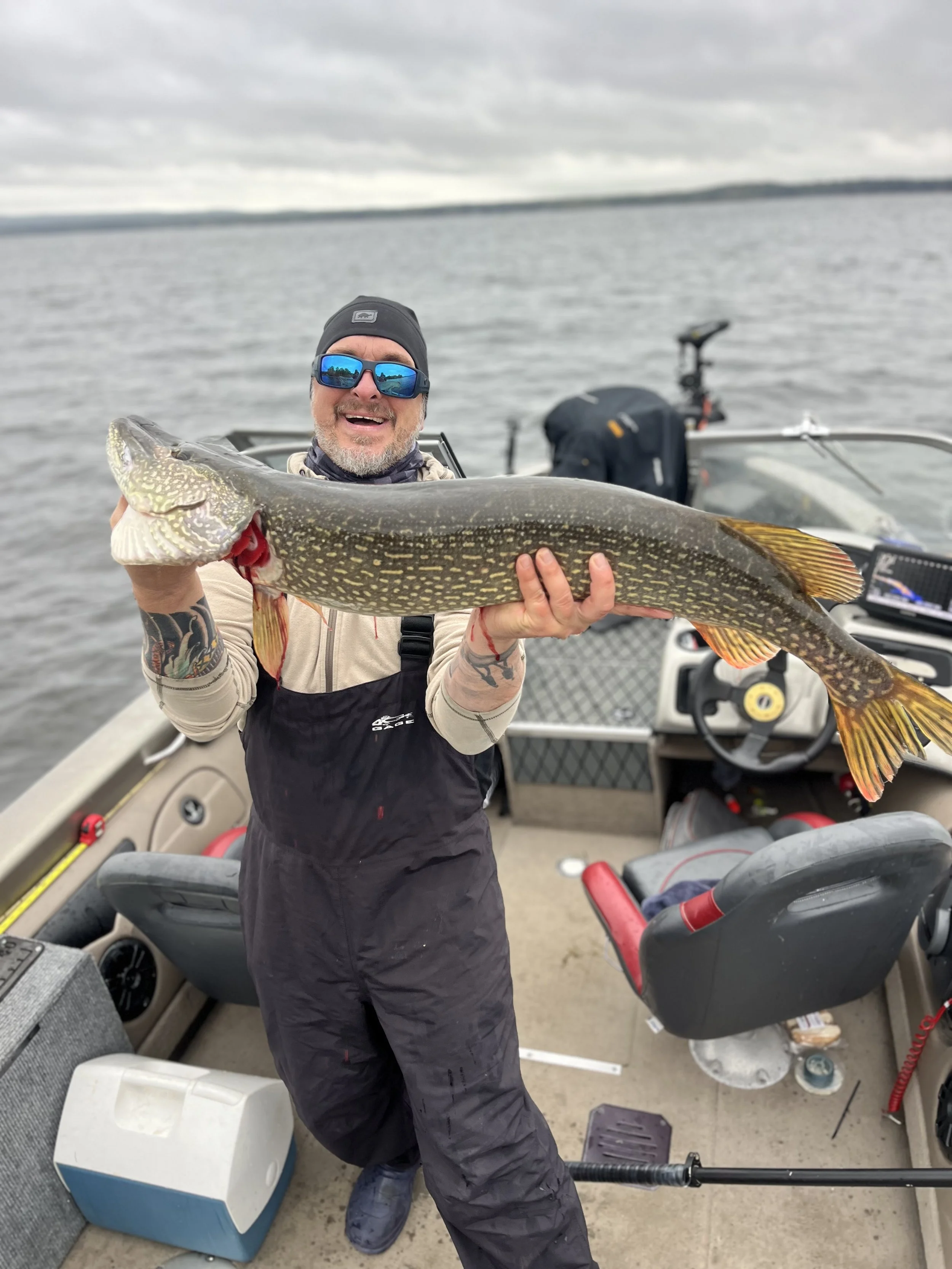 Man in sunglasses holding a large fish on a boat on a lake, with cloudy sky in the background.