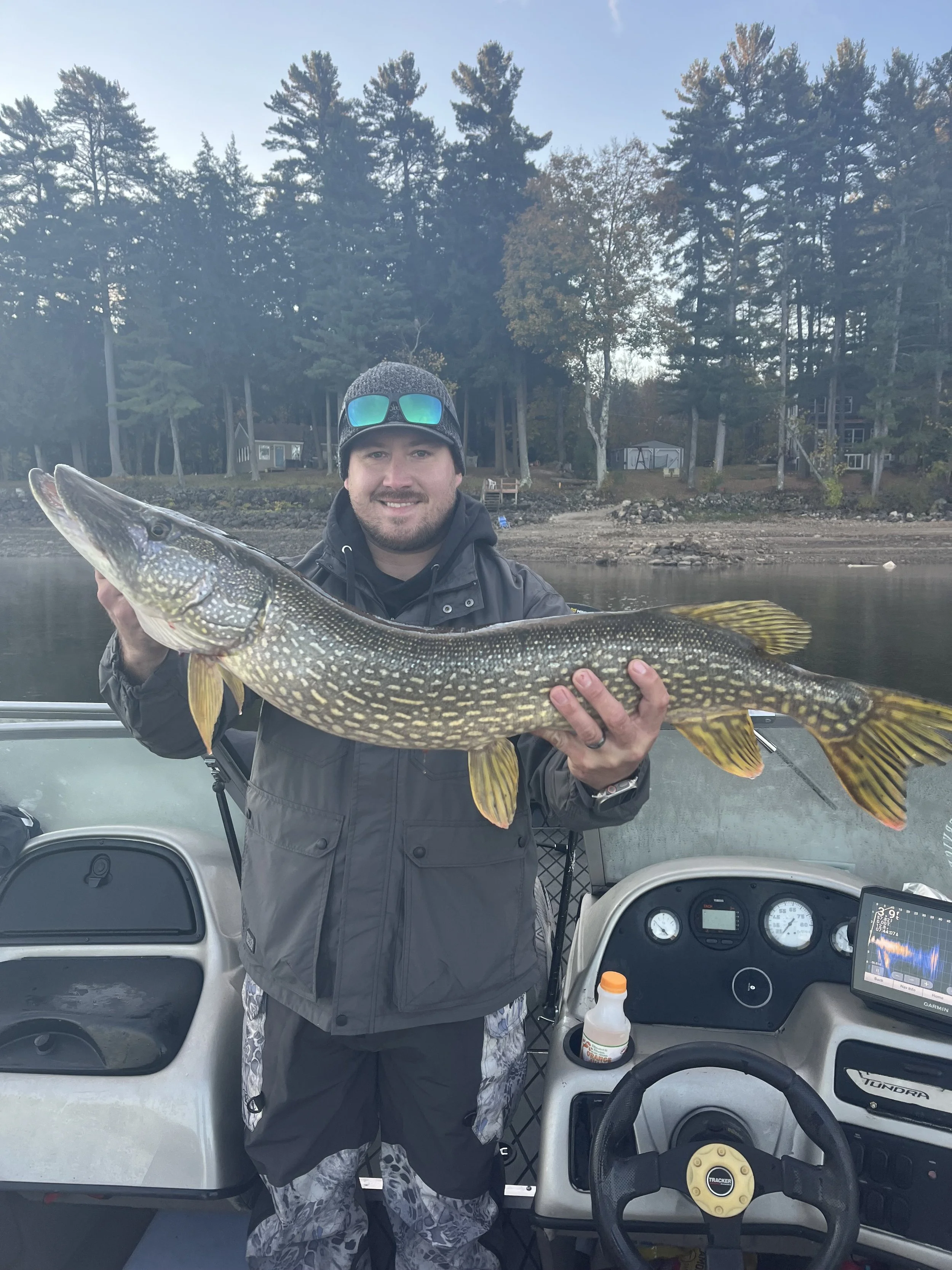 Man holding a large fish on a boat with a lake and wooded shoreline in the background.