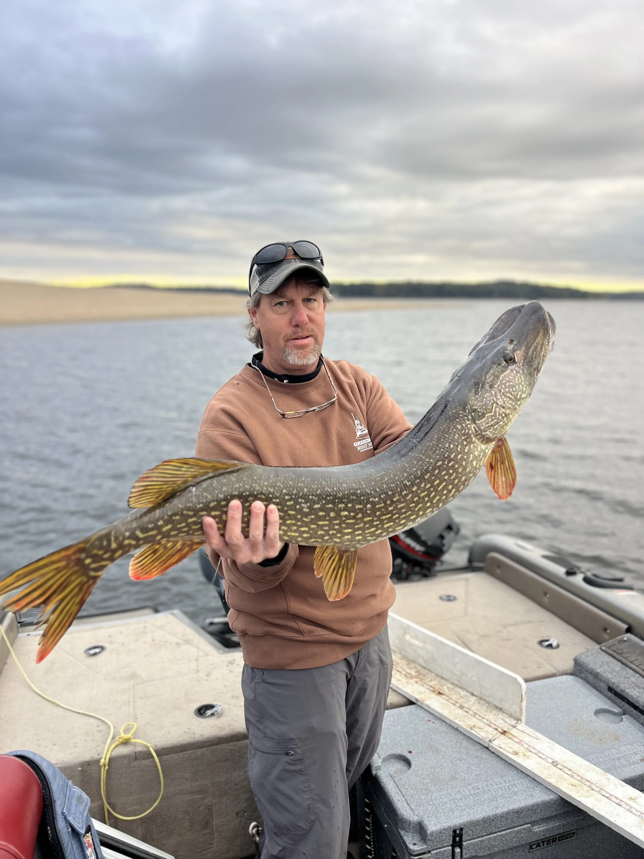 Man holding a large fish on a boat on a lake with cloudy sky.