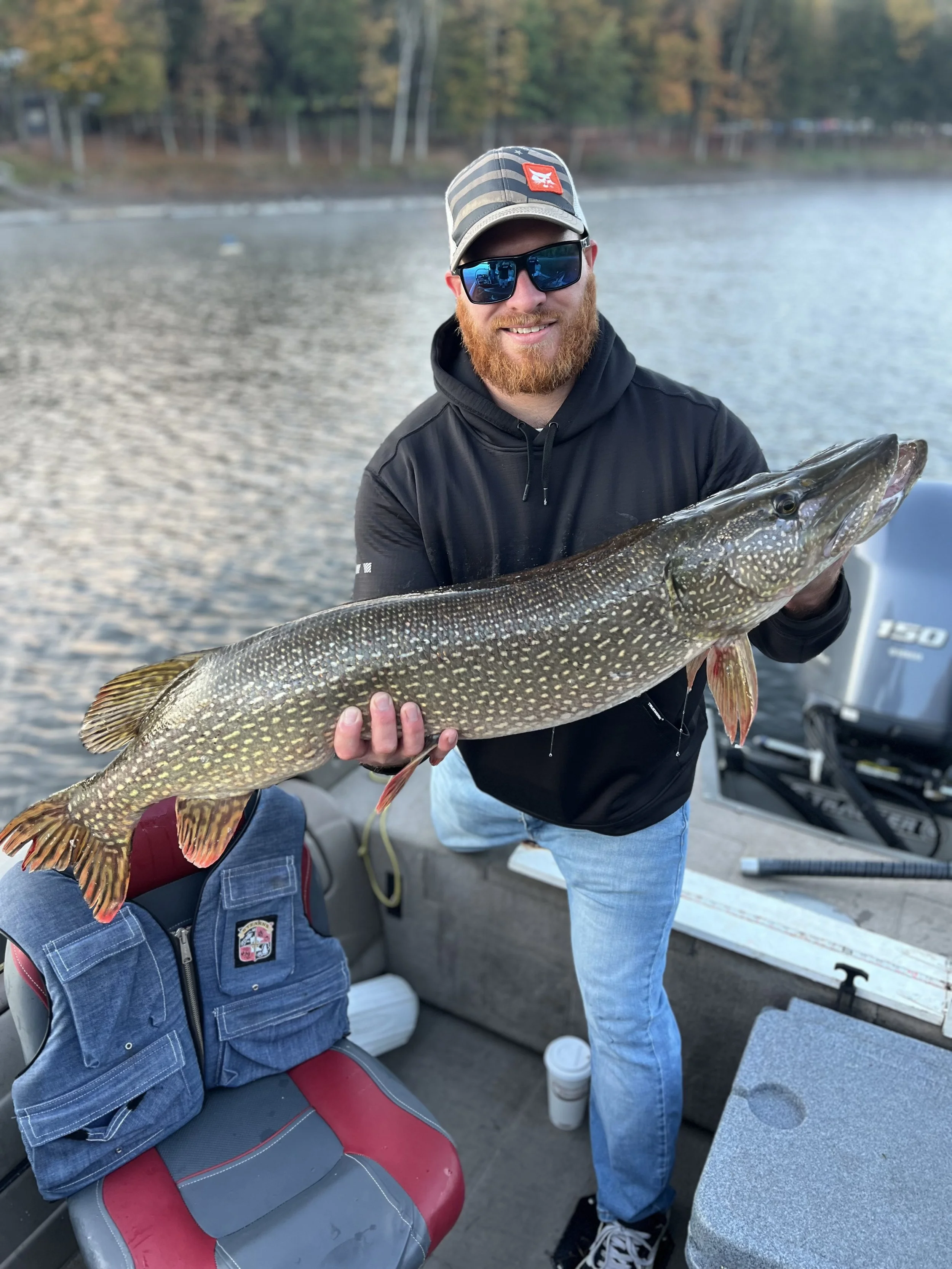 A man with a beard, wearing sunglasses, a baseball cap, and a black hoodie, smiling and holding a large fish, likely a northern pike, on a boat by a lake, with trees in the background.