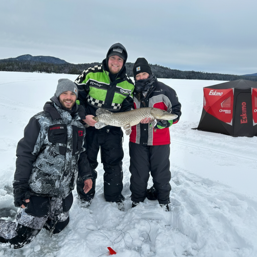 Man standing on snow-covered ice holding a large fish outdoors with trees in the background.