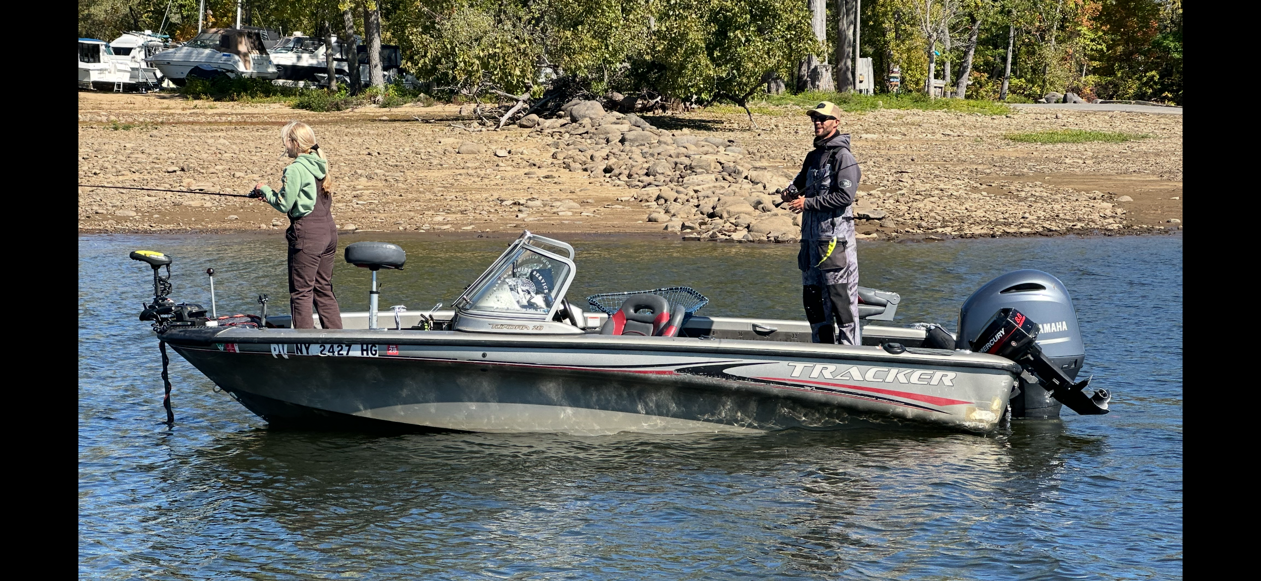 A small Tracker boat on a trailer parked near a body of water with hills and trees in the background and a partly cloudy sky.