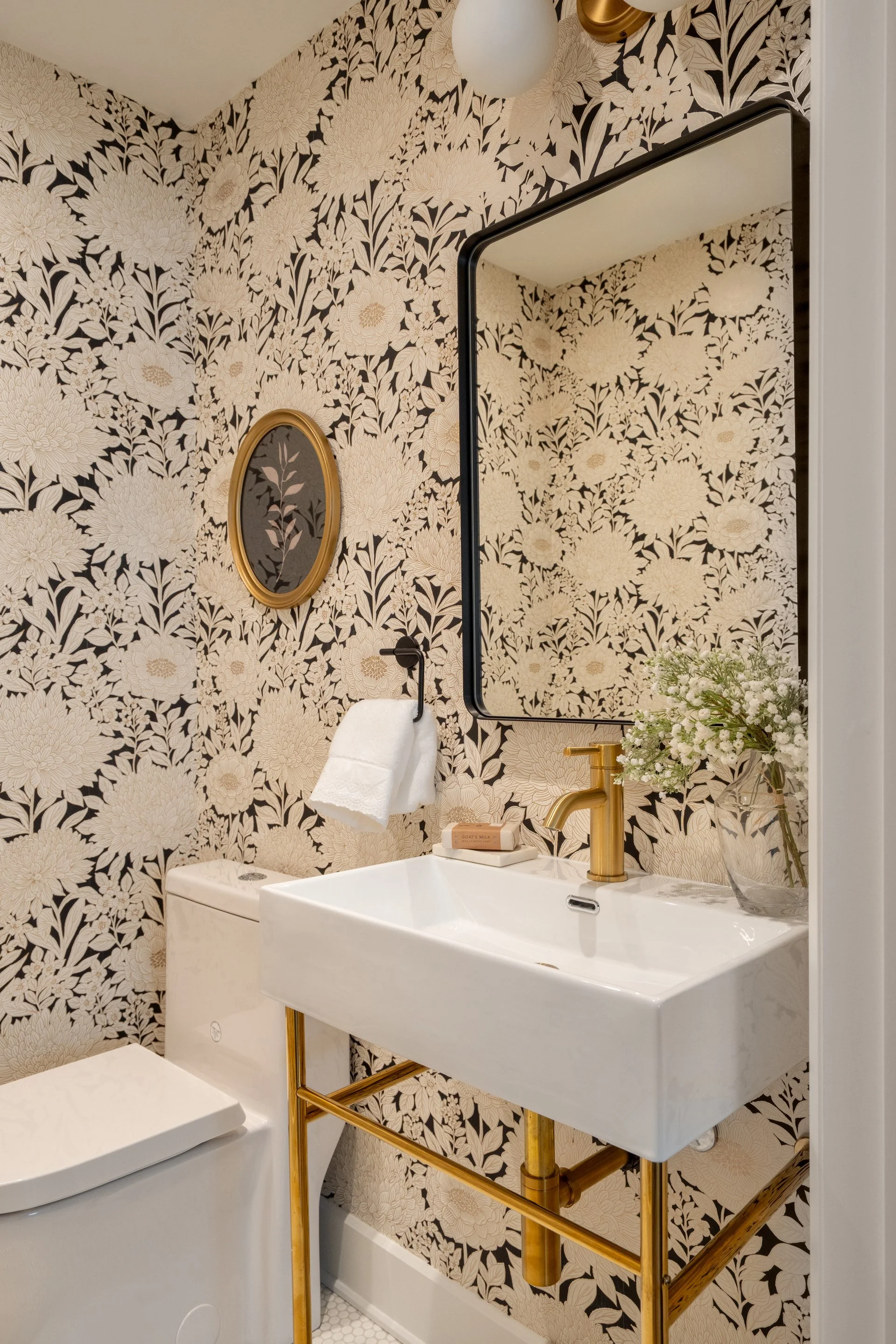 A small bathroom with floral wallpaper, a white sink with gold fixtures, a gold-framed mirror, and a small oval mirror on the wall. There is a vase with white flowers on the sink and a towel hanging on a black hook.