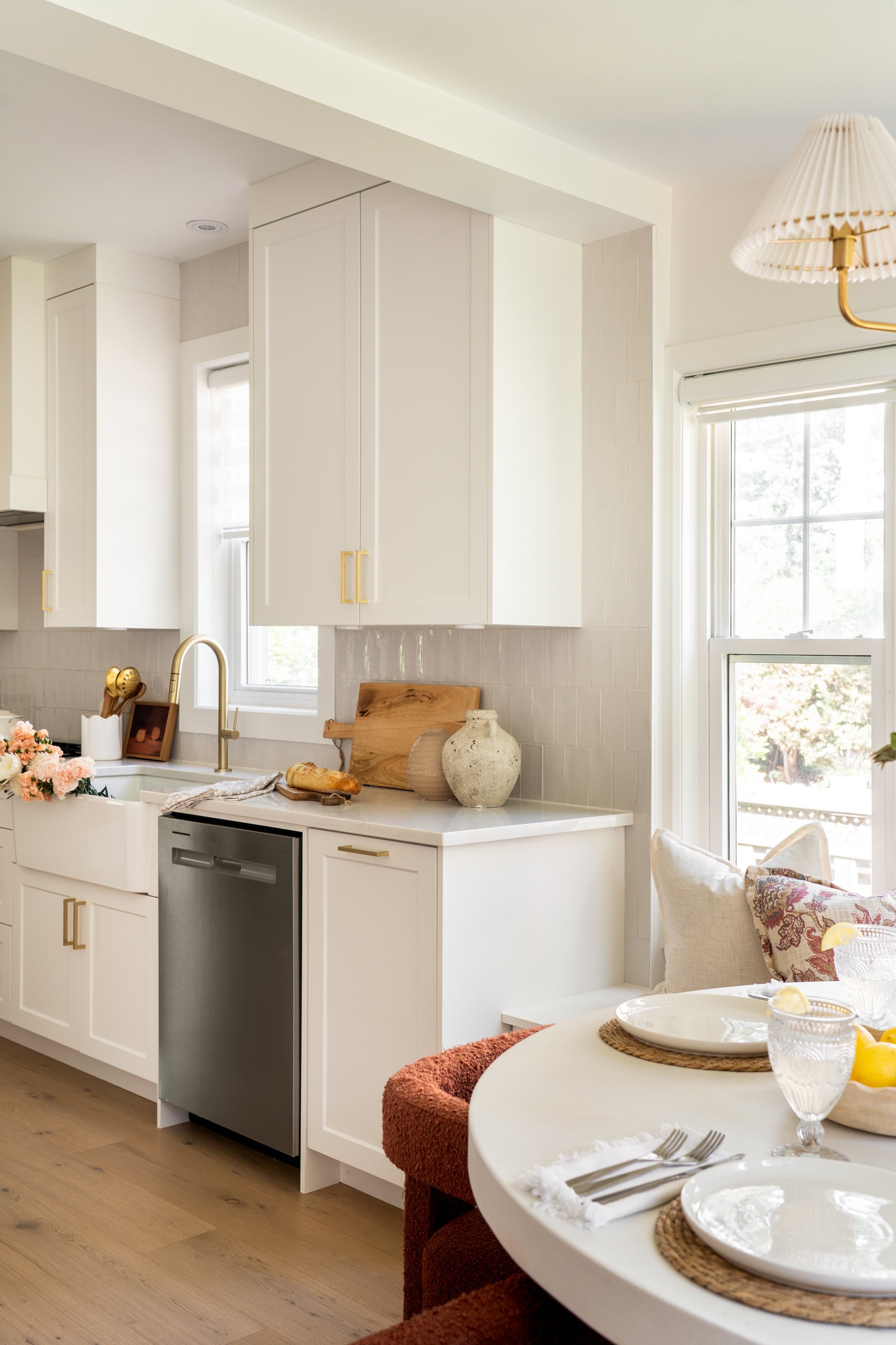 A bright, modern kitchen with white cabinets, a gold faucet, a window above the sink, and a small dishwasher. To the right, there is a dining table set with plates, glasses, and lemons, with chairs covered in reddish-brown fabric. Decor items like vases, a wooden cutting board, and flowers are on the countertop.