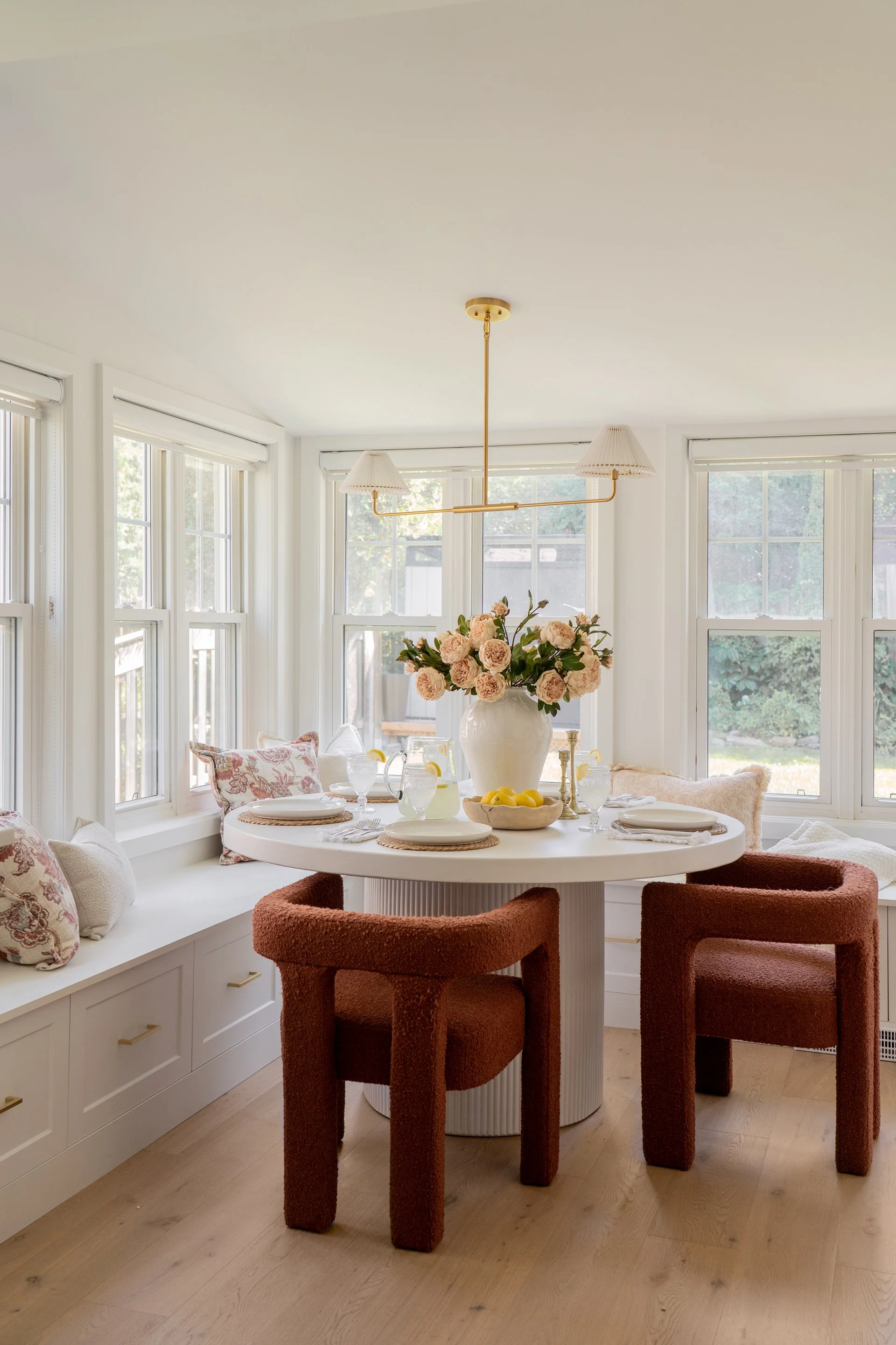 A bright dining nook with a round white table, surrounded by two rust-colored upholstered chairs. Window seat with cushions and pillows runs along the wall with multiple windows allowing natural light. The table is set with white dishes, glassware, and a large white vase of pink roses. A gold ceiling light with two lampshades hangs overhead.