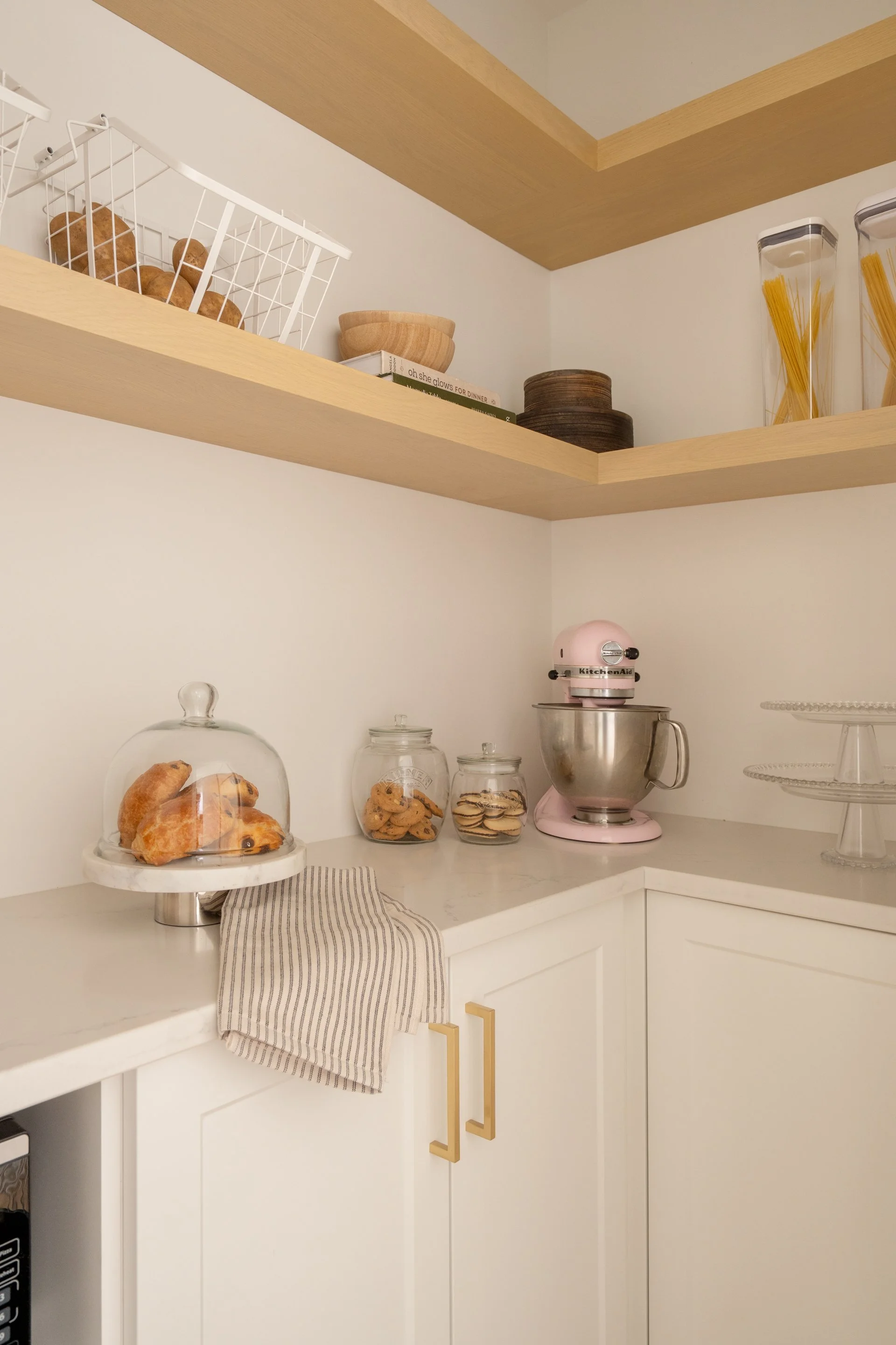 Kitchen countertop with a cake stand holding croissants, three glass jars with cookies, a pink KitchenAid stand mixer, and a two-tier glass cake stand. Shelves above with bowls, cookbooks, pottery, and pasta jars.