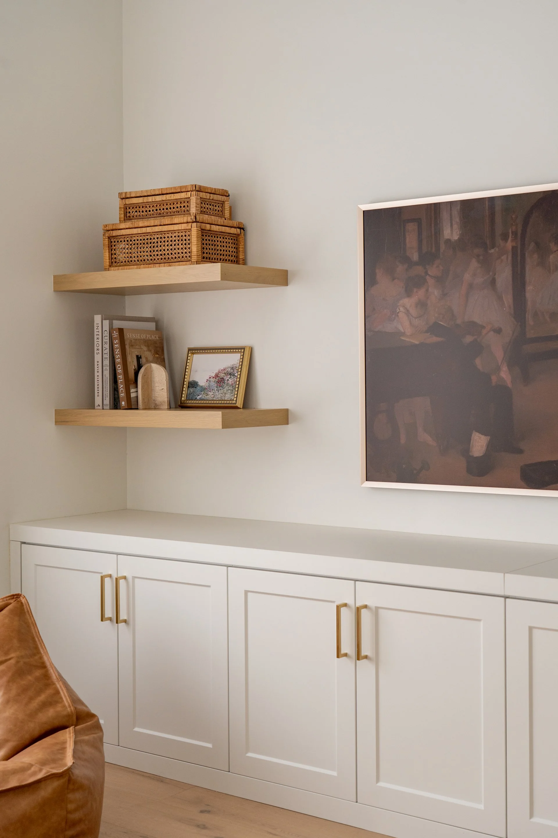 Decorative white cabinet with gold handles, two wooden shelves with wicker baskets, books, a small framed painting, a decorative stone, and a large framed painting on a white wall.