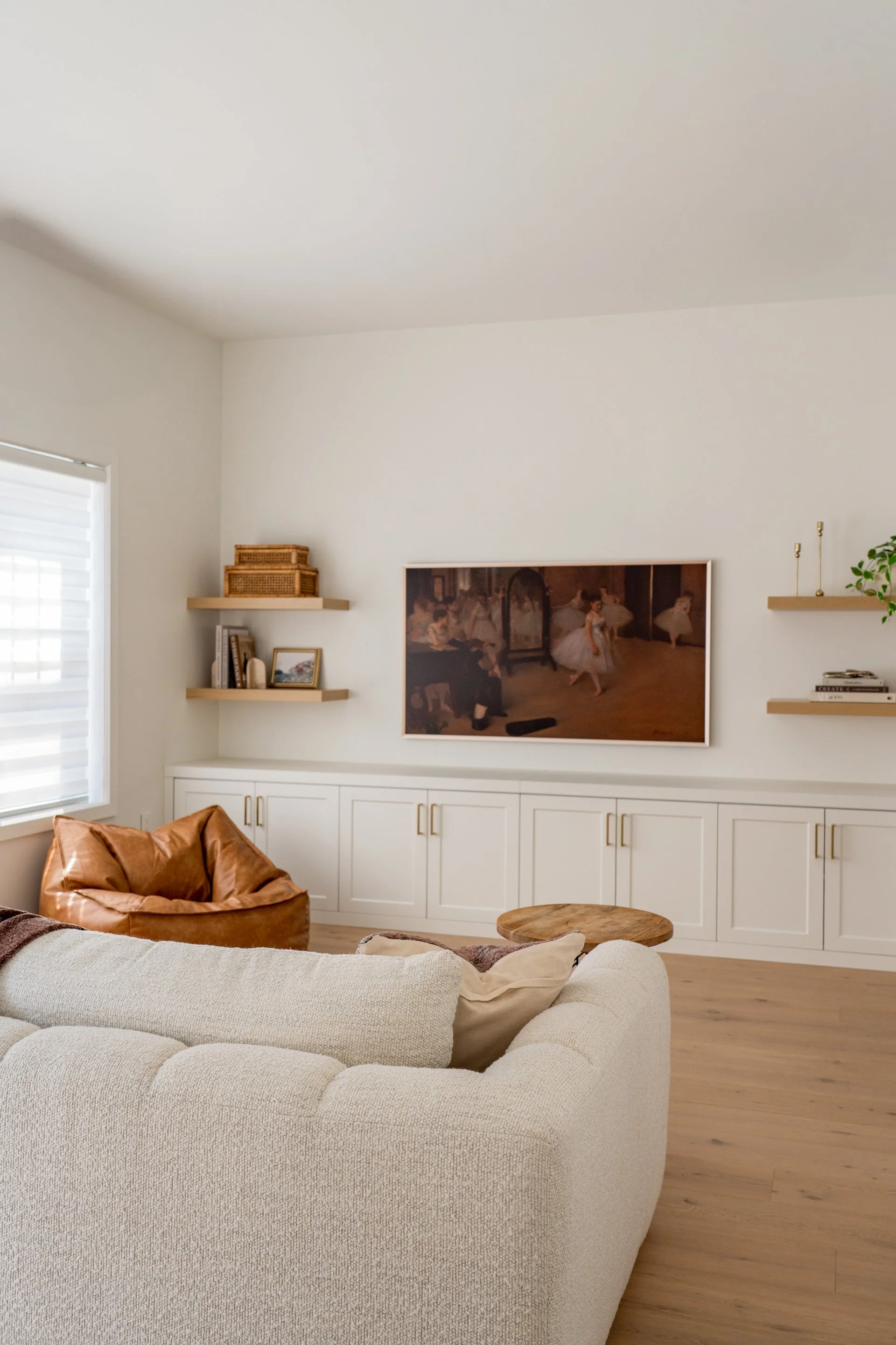 A cozy living room with a cream-colored sectional sofa, a brown leather chair, and a round wooden side table. The room has white walls, built-in white cabinetry, and floating shelves with decor items and books. A flat-screen TV displays a ballet performance scene. A window with blinds lets in natural light.