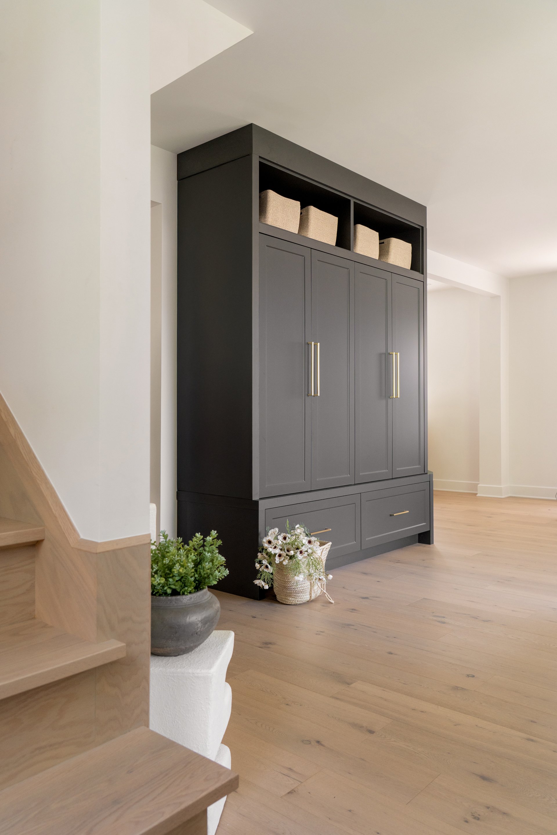 A dark gray built-in cabinet with gold handles, open shelves with beige baskets, on a light wood floor in a modern, bright living space.