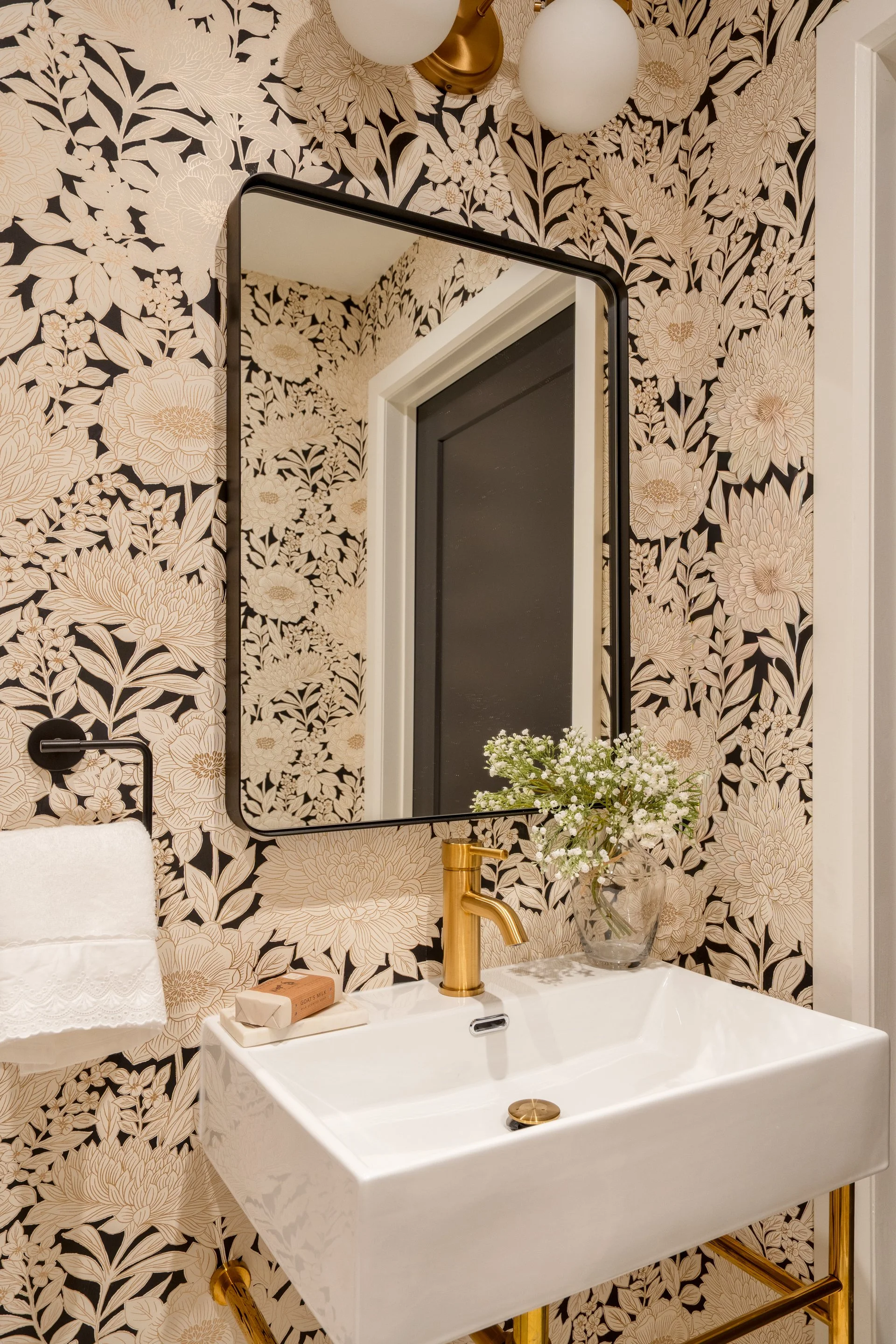 Decorative bathroom with floral wallpaper, a mirror, a white sink with brass fixtures, a soap bar, a small glass vase with white flowers, and spherical wall lights.