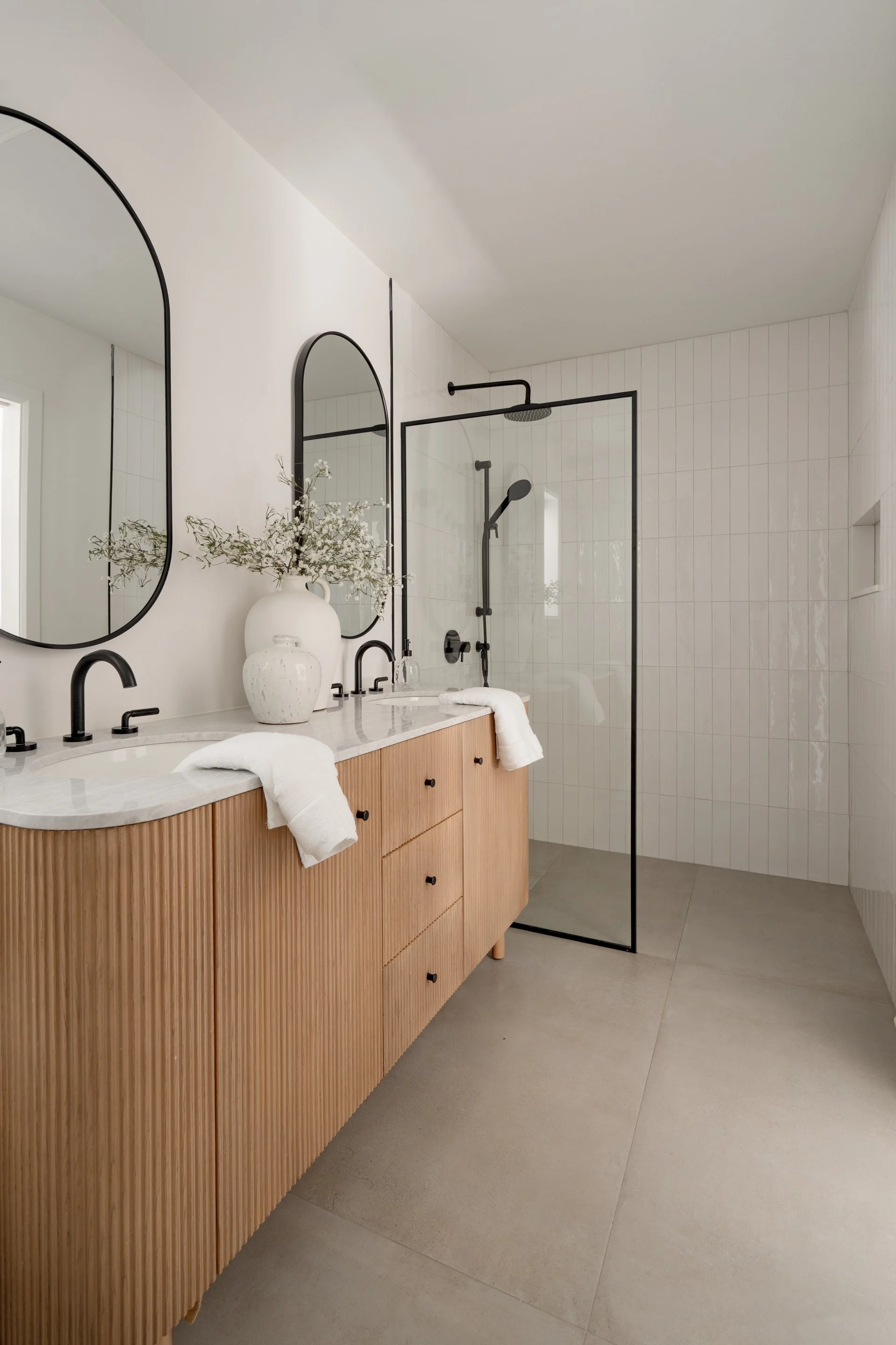 Modern bathroom with dual sinks, black fixtures, two oval mirrors, wooden vanity, ceramic vases with decorative branches, and a glass-enclosed walk-in shower with black hardware and white tiled walls.